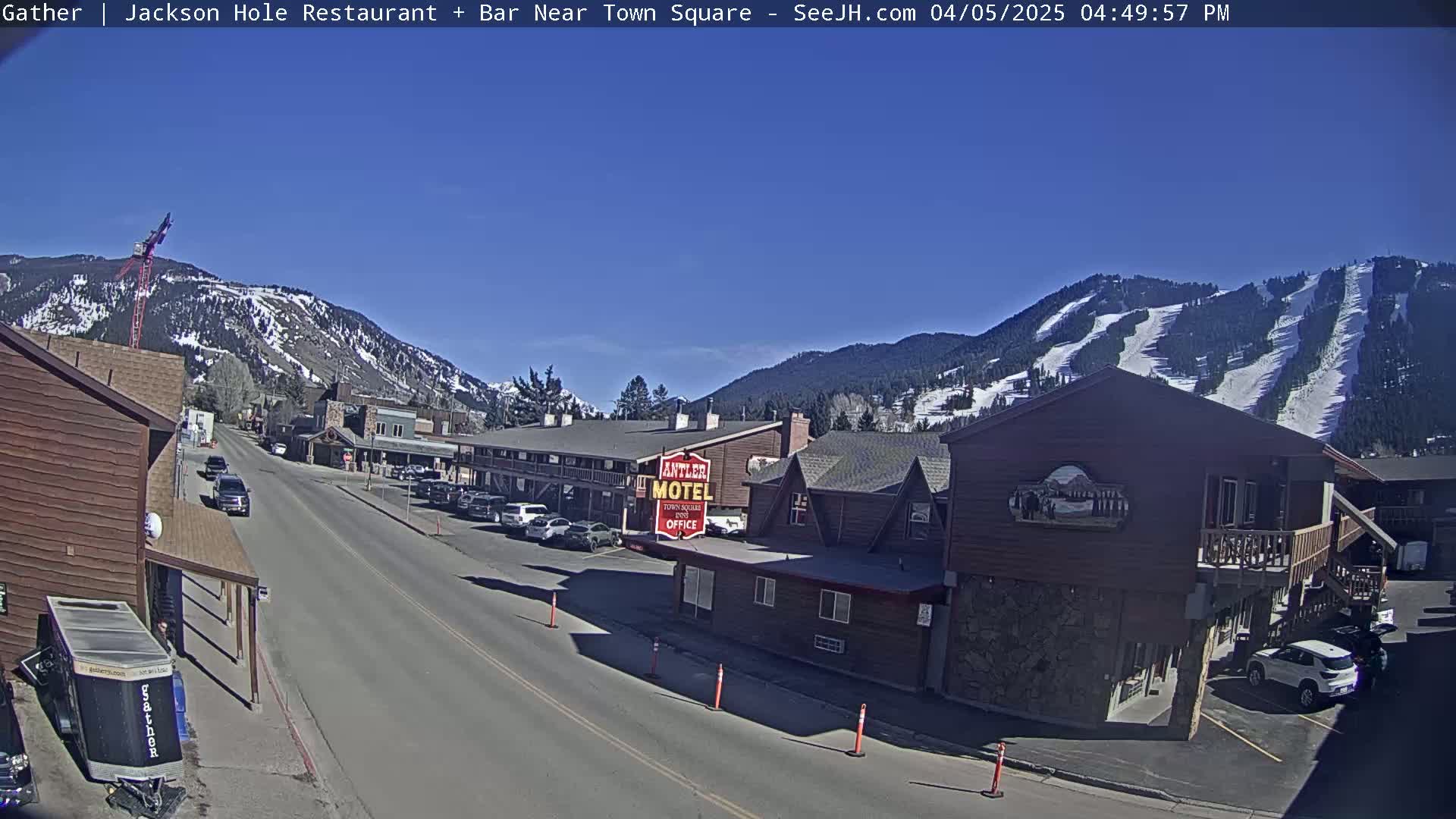 A sunny day reveals a town street scene nestled between snow-capped mountains with ski runs visible on one, featuring several buildings including a motel, cars parked along the sides of the road, and a construction crane in the distance.