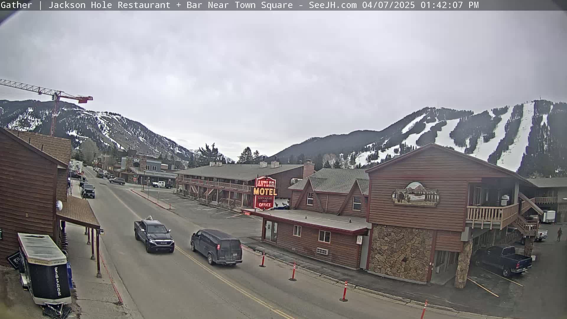 A street scene shows a road with several vehicles, buildings, including a motel, and a mountain range in the background with ski slopes visible under an overcast sky.