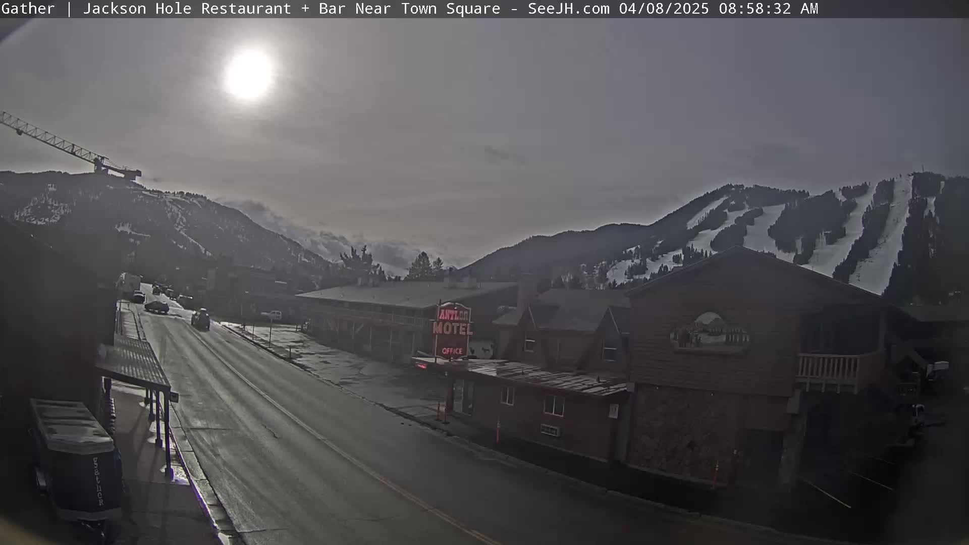 A wet street with cars driving past a motel, under a hazy sun, with snow-covered mountains in the background.