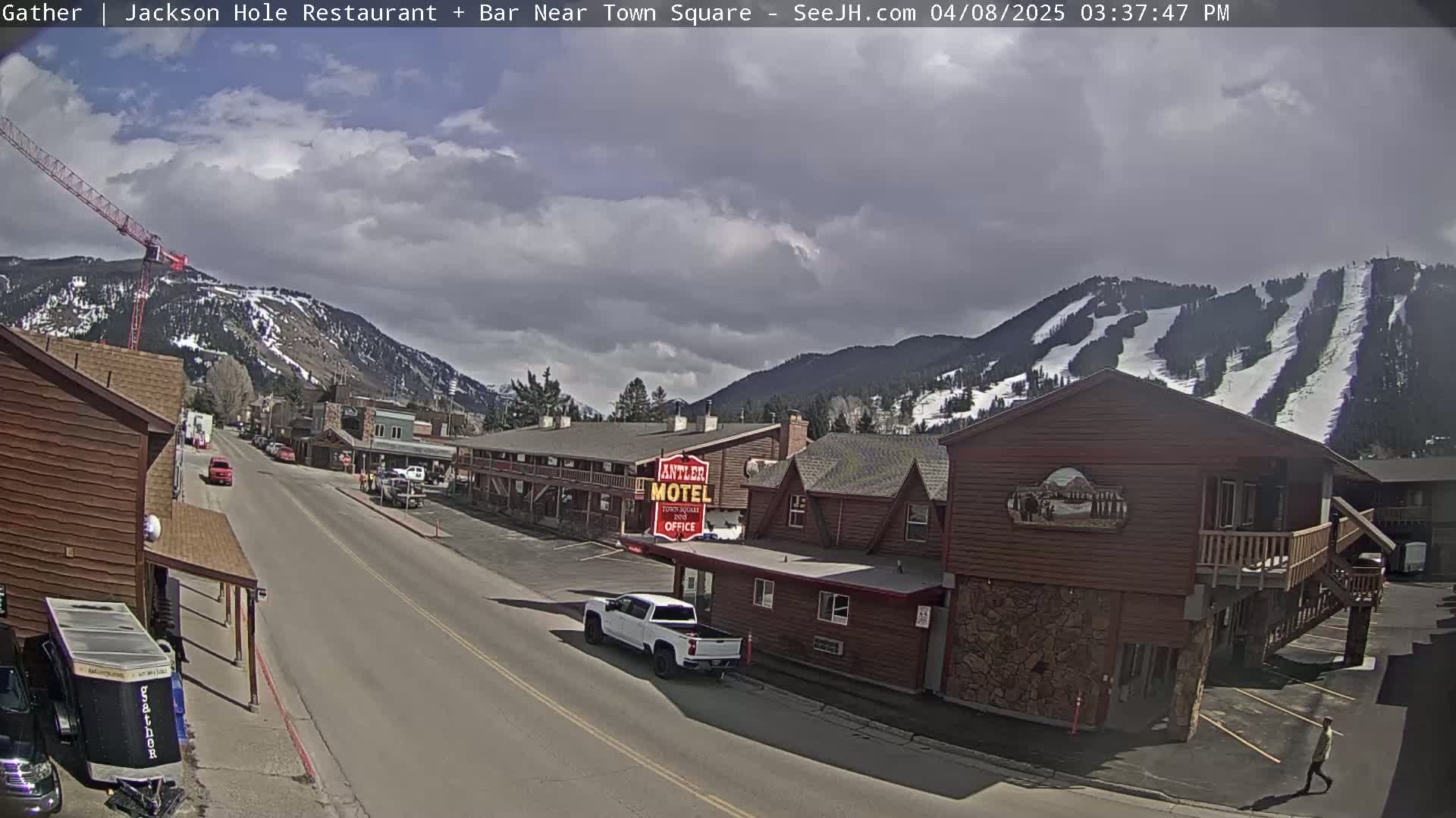 A partly cloudy day reveals a street scene in a mountain town, with snow-covered slopes visible in the background.