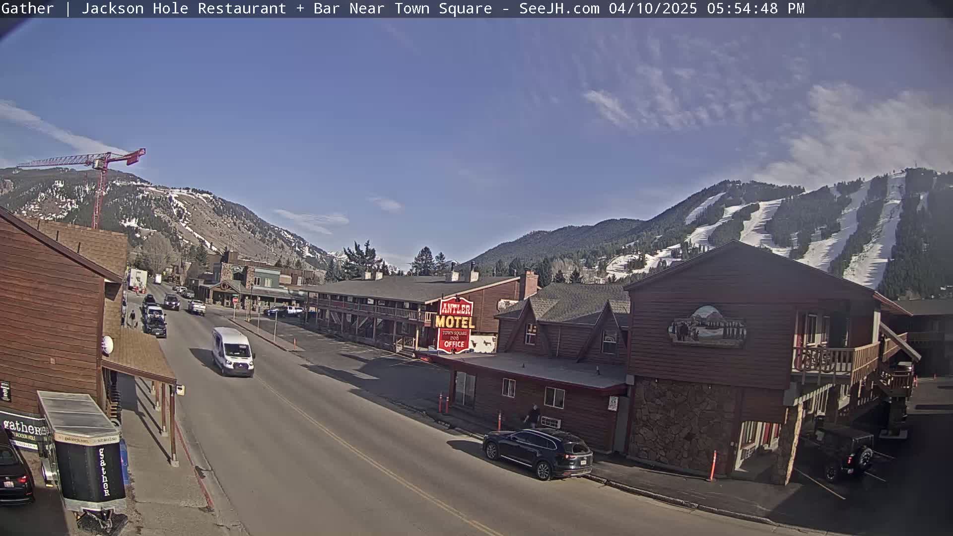 A sunny day reveals a street scene in a mountain town, with cars parked and driving along a road lined with buildings, including a motel, against a backdrop of snow-capped mountains.
