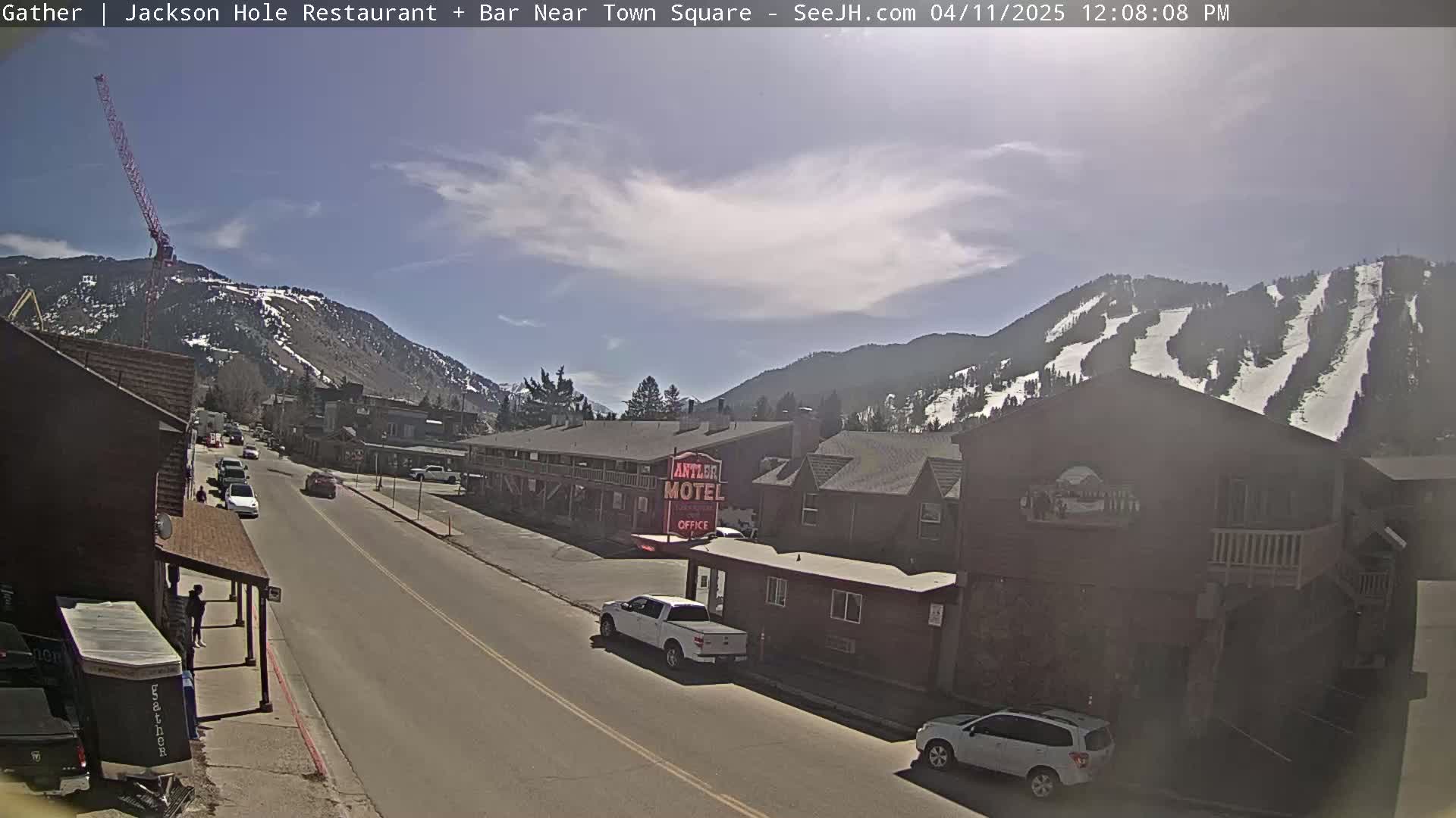 A sunny day reveals a street scene in a mountain town with vehicles parked along the road, buildings lining the street, a motel, and a mountain with ski runs in the background, under a partly cloudy sky.