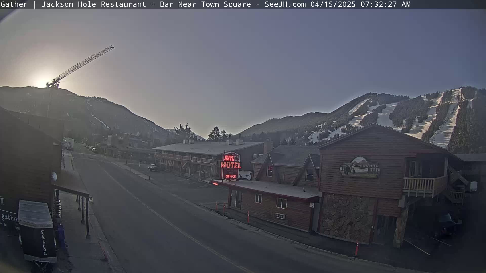A street scene in a mountain town at sunrise shows a motel, a crane on a hillside, and a road leading toward snow-covered mountains.