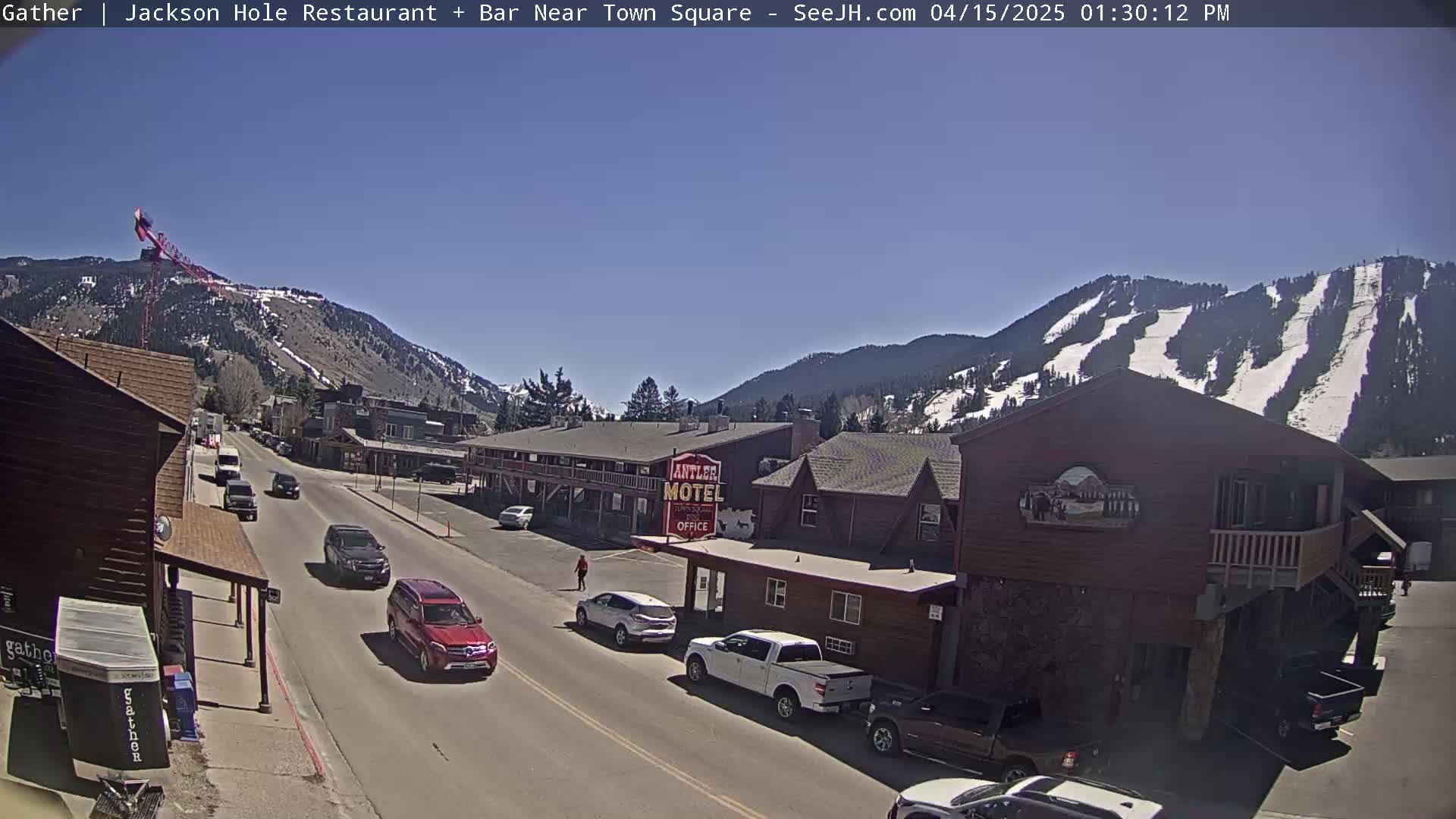 A sunny day reveals a street scene in a mountain town, with vehicles parked and driving past a motel and other buildings, all set against a backdrop of snow-capped mountains.