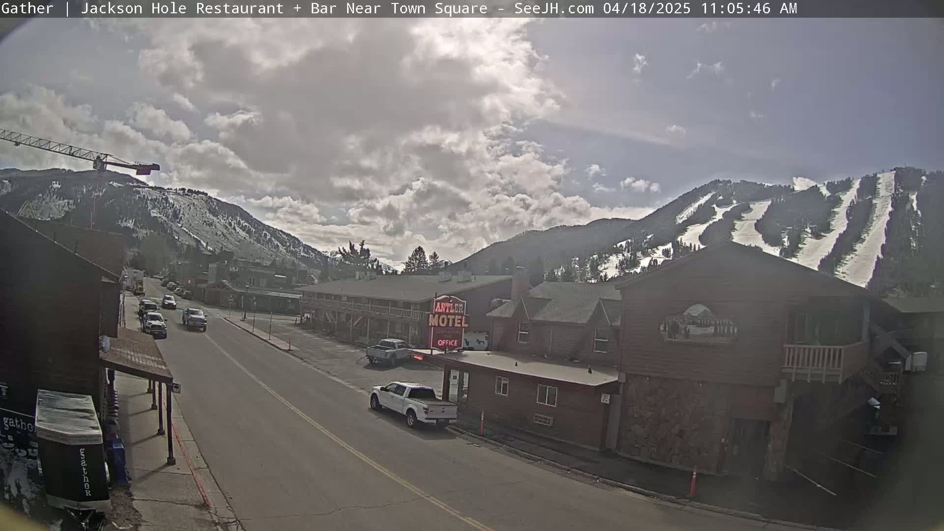 A partly cloudy day reveals a street scene with cars driving past a motel and buildings, mountains with ski runs in the background, and a construction crane visible to the left.