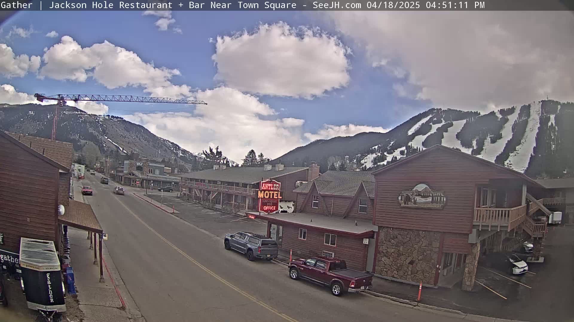 A partly cloudy day shows a town street scene with cars parked and driving past buildings, a construction crane, and a snow-covered mountain in the background.