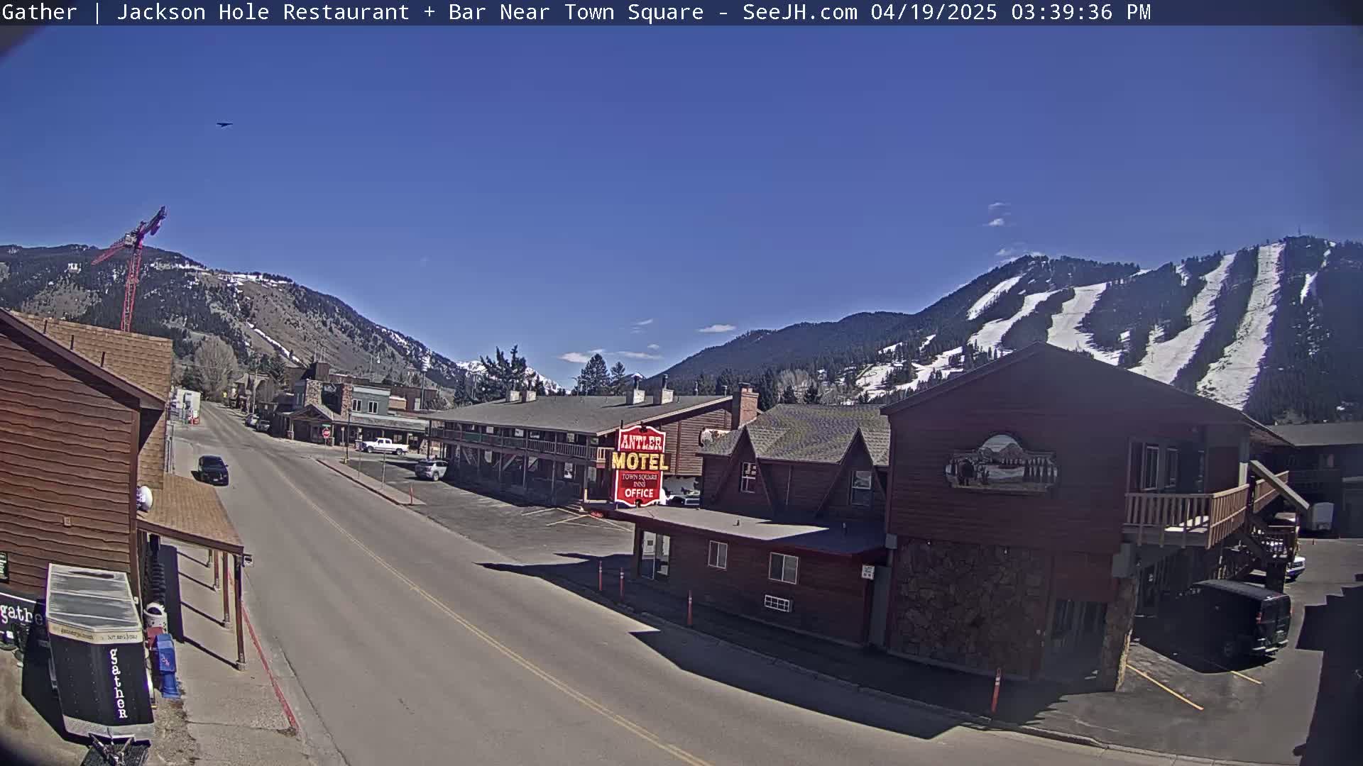 A mostly sunny day reveals a street scene in a mountain town, with buildings lining a road that leads toward snow-covered mountains with visible ski runs.