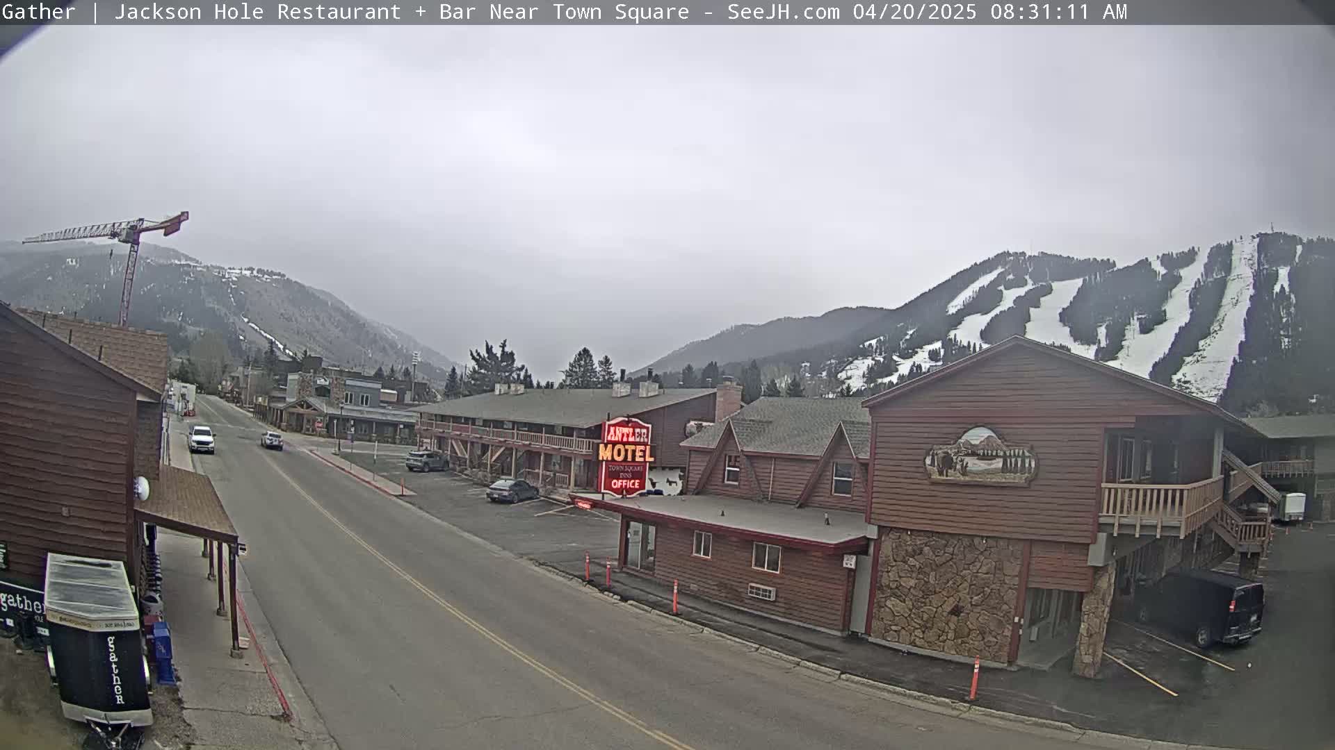 A street scene on an overcast day shows a road lined with buildings, including a motel, with a mountain range in the background showing ski slopes.