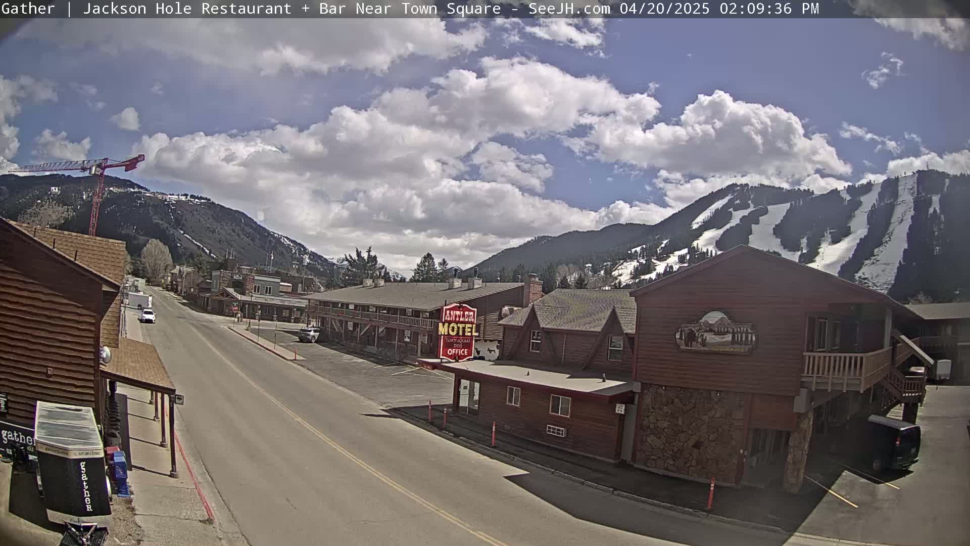 A partly cloudy day reveals a street scene in a mountain town with a motel, buildings, and a distant ski slope.