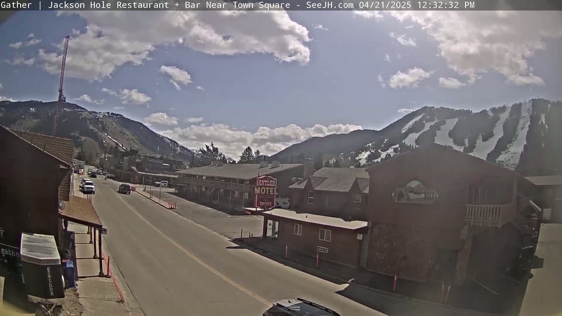 A partly sunny day reveals a street scene in a mountain town, with buildings, cars, and snow-covered mountains in the background.
