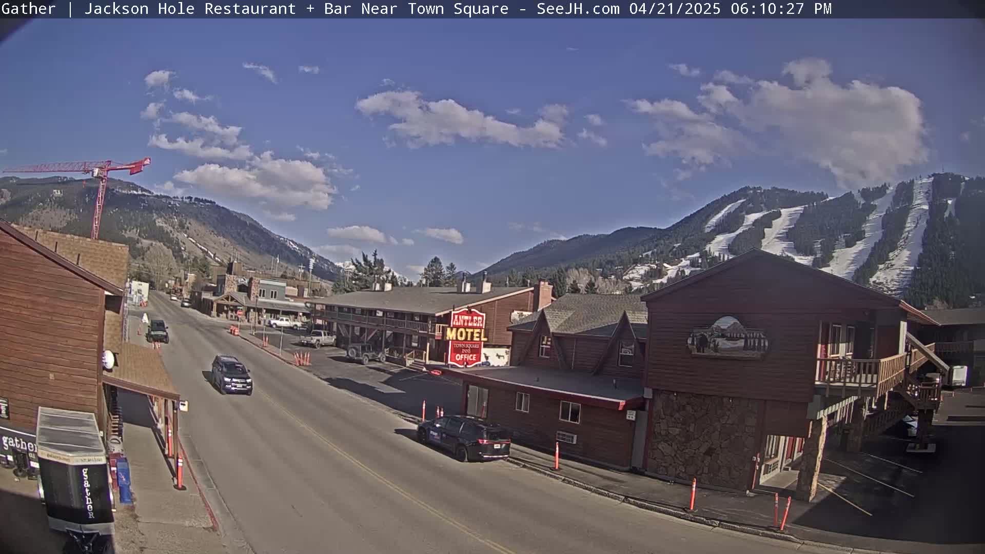 A partly sunny day reveals a mountain town street scene with a motel, cars, and a construction crane, all set against a backdrop of snow-covered mountains with visible ski runs.