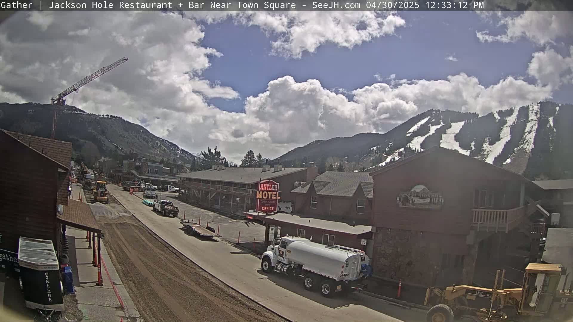 A partly cloudy day reveals a street scene in a mountainous area with construction underway, featuring buildings, vehicles, and a crane against a backdrop of snow-capped hills.