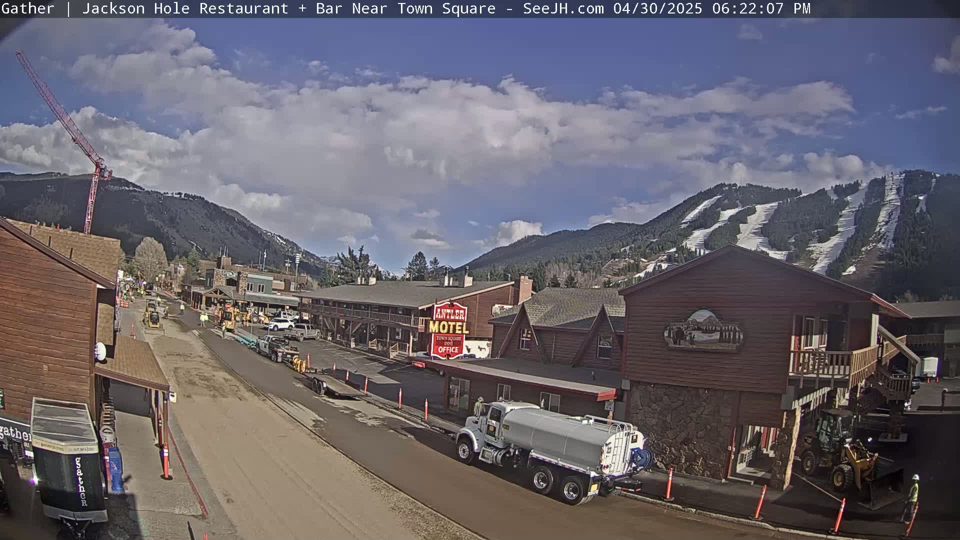 A partly cloudy day reveals a town street scene with construction equipment, a water truck, and a motel, all set against a backdrop of snow-capped mountains.