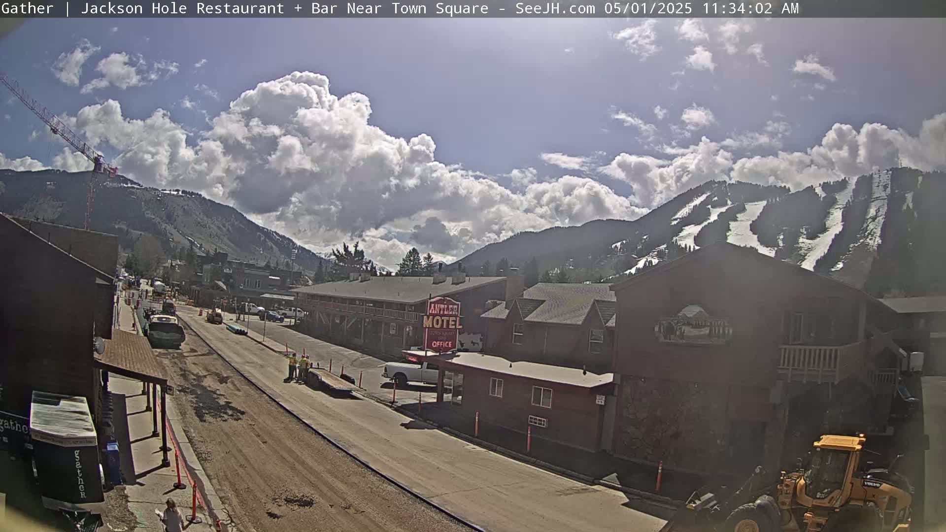A partly cloudy day reveals a street scene in a mountain town with a motel, construction equipment, and snow-covered mountains in the background.