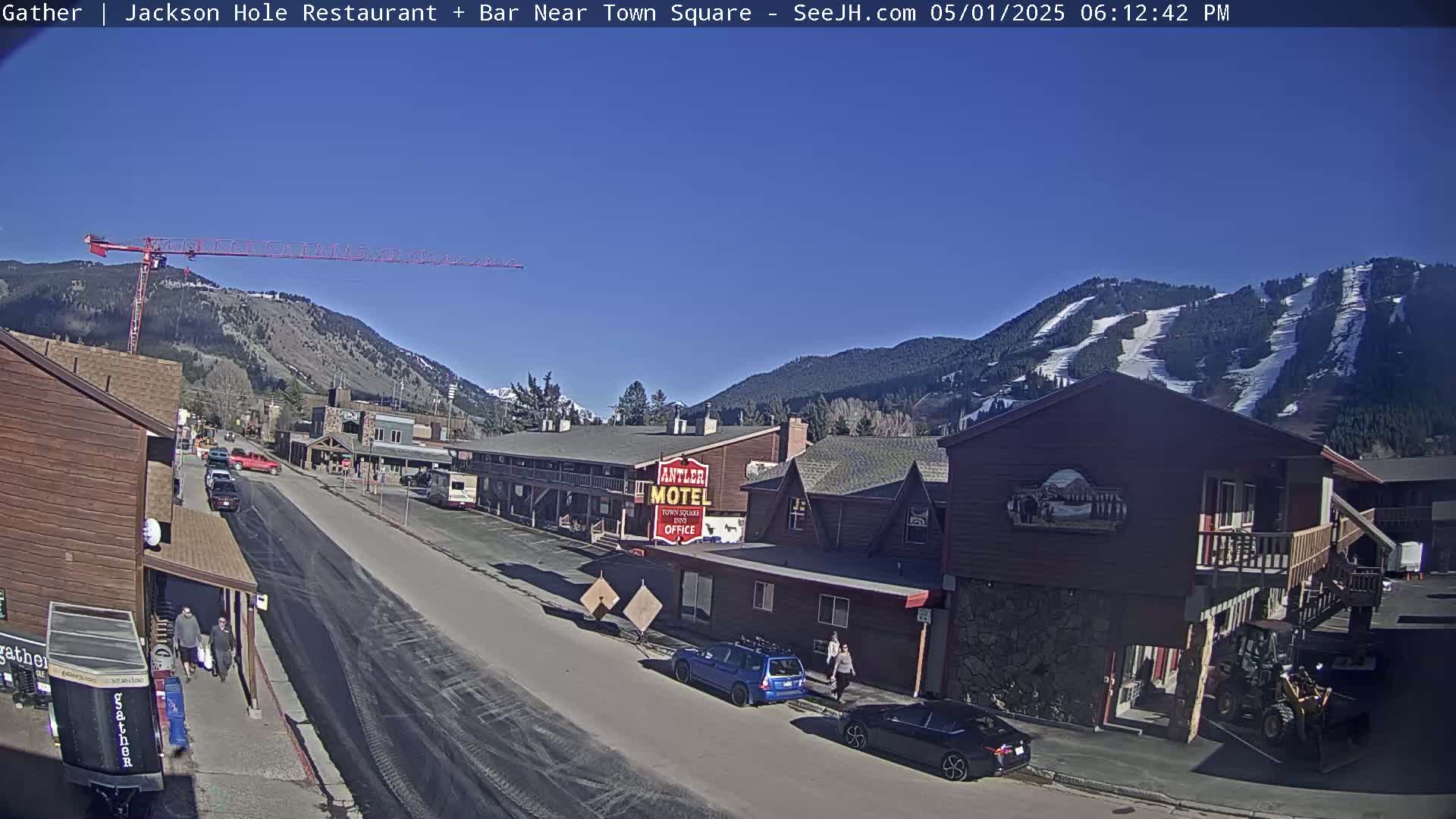 A sunny day reveals a town street scene with buildings, parked cars, and a construction crane against a backdrop of snow-capped mountains.