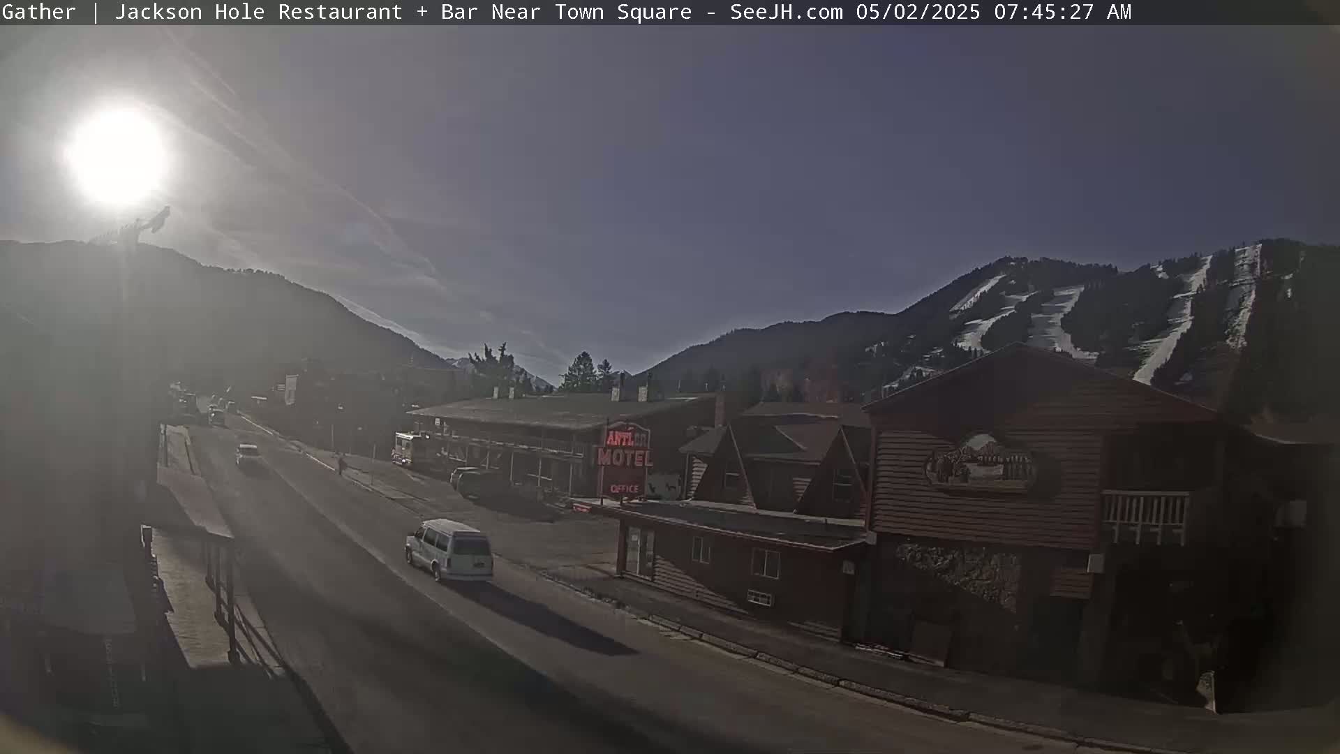 A sunny morning view of a town street with cars and buildings, including a motel, nestled in a valley with a mountain slope showing ski runs in the background.
