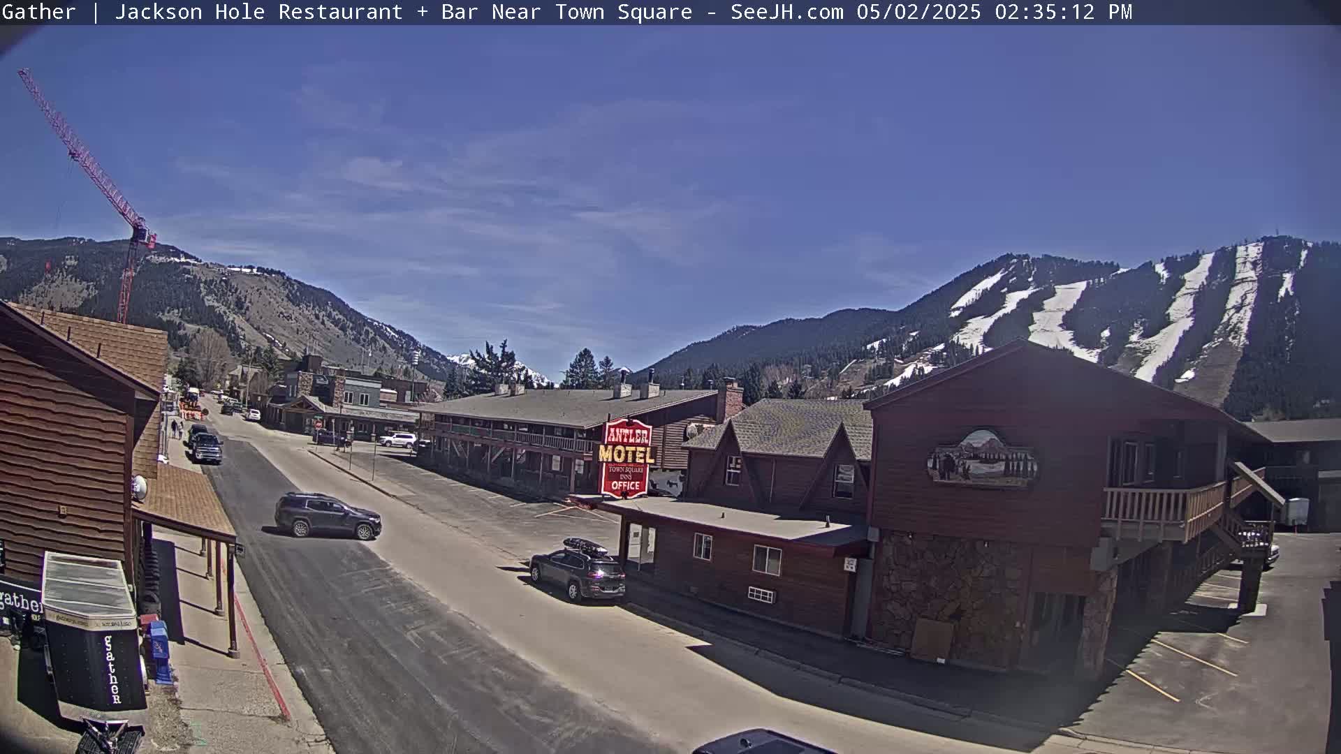 A sunny day reveals a street scene in a mountain town with a motel, cars, and a construction crane visible against a backdrop of snow-covered mountains.