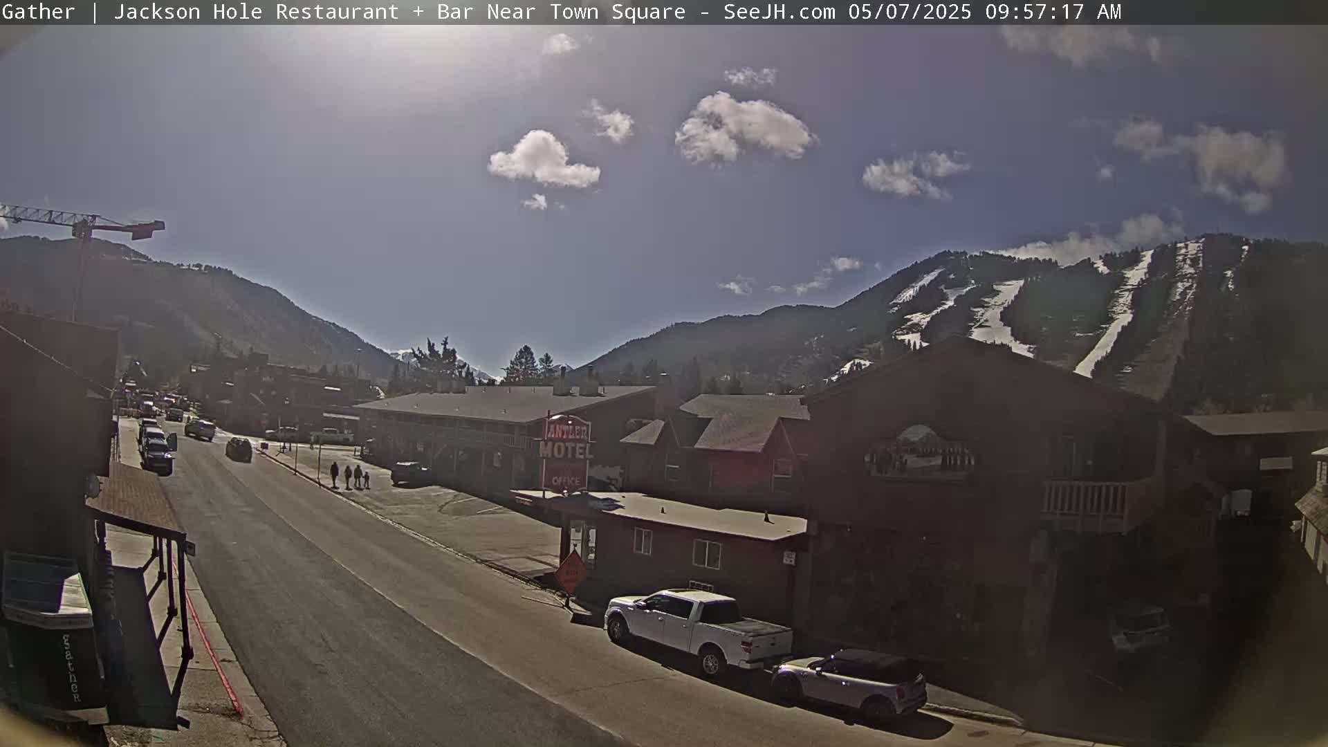 A sunny day in a mountain town shows a street scene with cars, pedestrians, and buildings, including a motel, with snow-covered mountains in the background.
