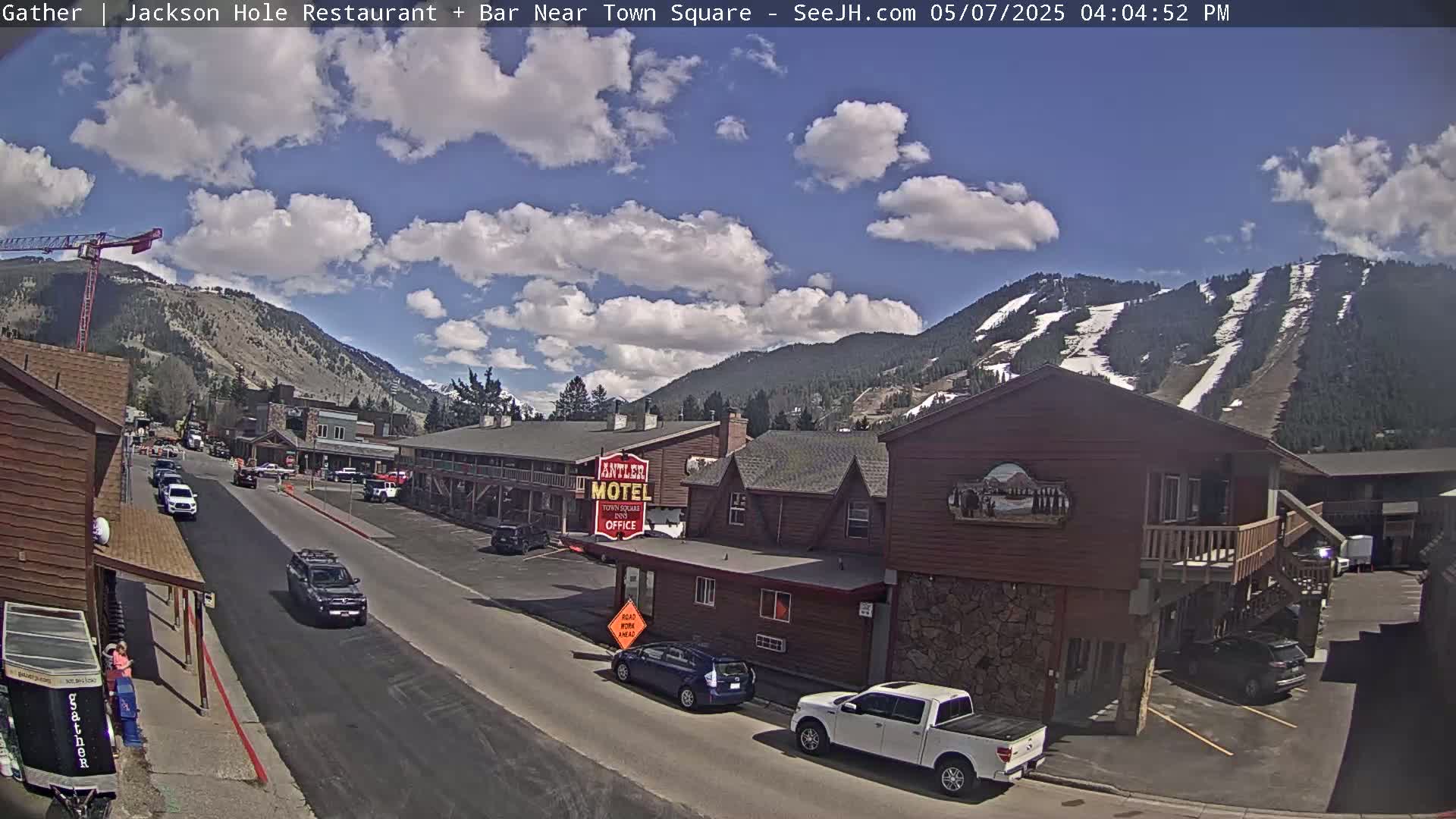 A partly sunny day reveals a street scene in a mountain town, with cars parked and driving past a motel and other buildings, and snow-covered mountains in the background.