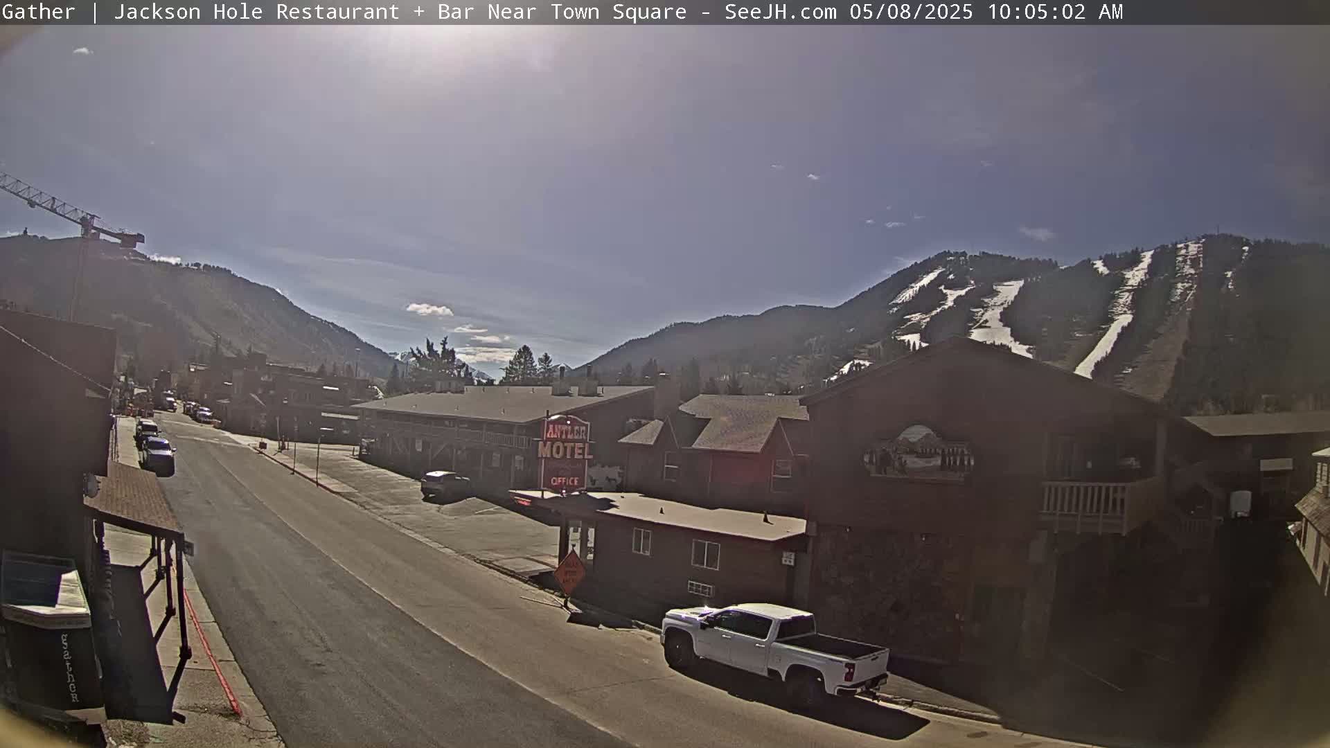 A sunny day reveals a town street scene nestled between mountains, one of which shows ski runs, with a motel and vehicles visible.