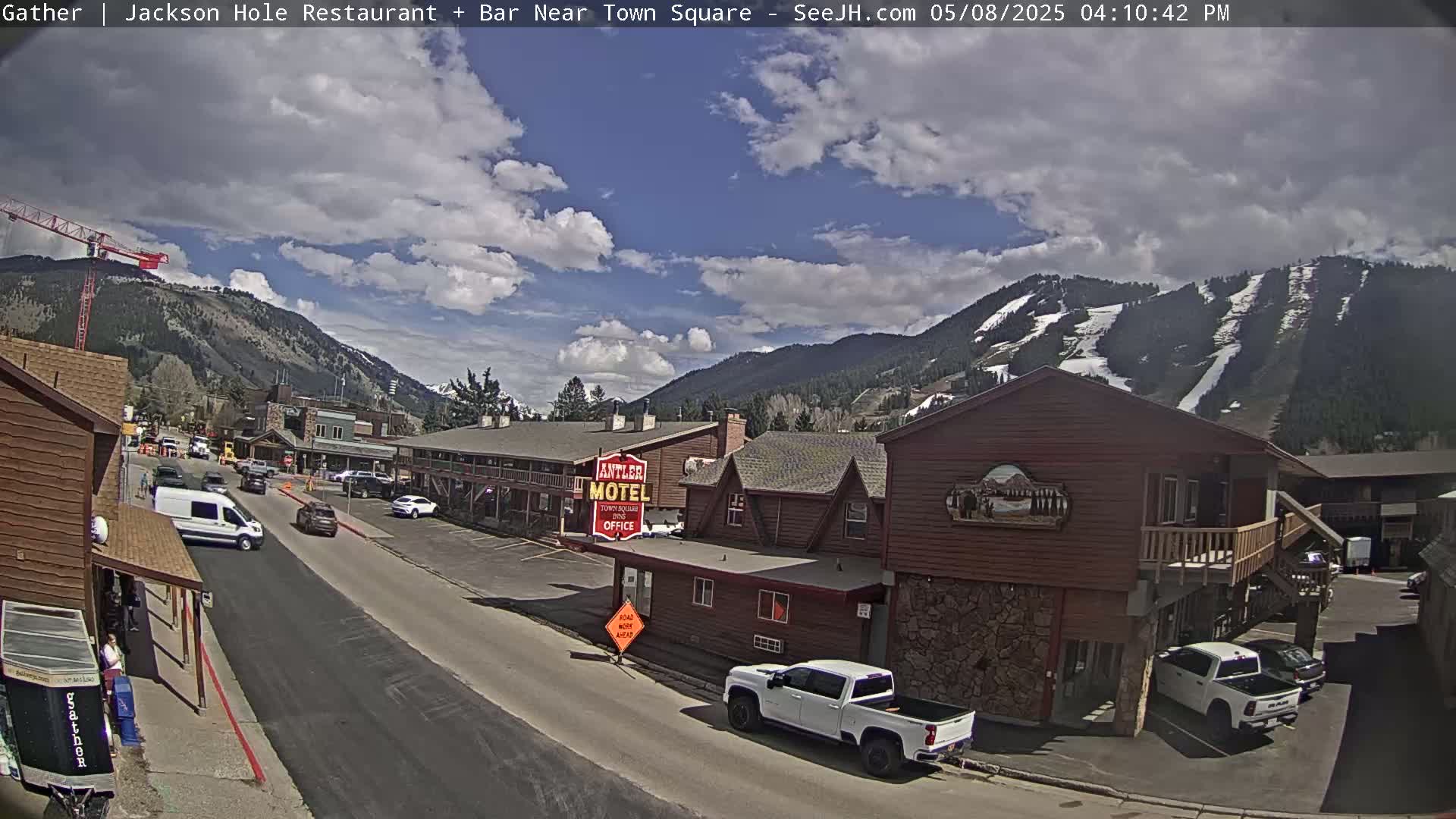 A partly cloudy day shows a street scene in a mountain town with cars parked along the roadside and a motel in view, with a mountain range in the background showing patches of snow.