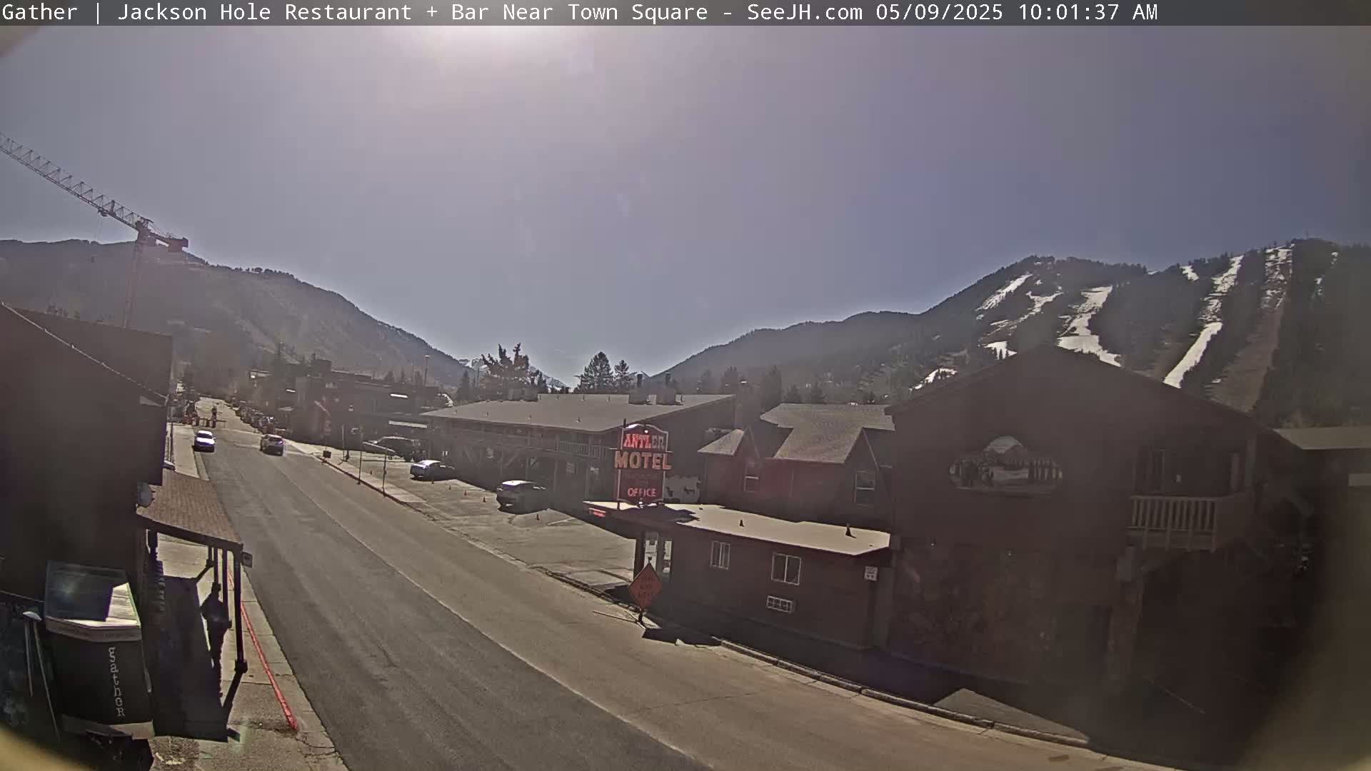 A sunny view of a street scene in a mountain town, with cars parked and driving along a road lined with buildings, including a motel, and snow-covered mountains in the background.