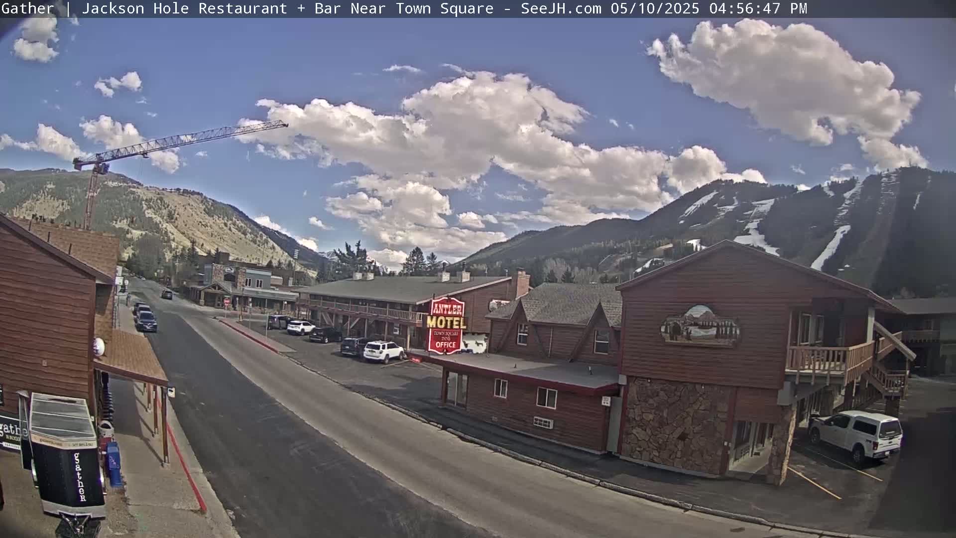 A partly cloudy day reveals a town street scene nestled in a valley between mountains, with a construction crane visible in the distance.