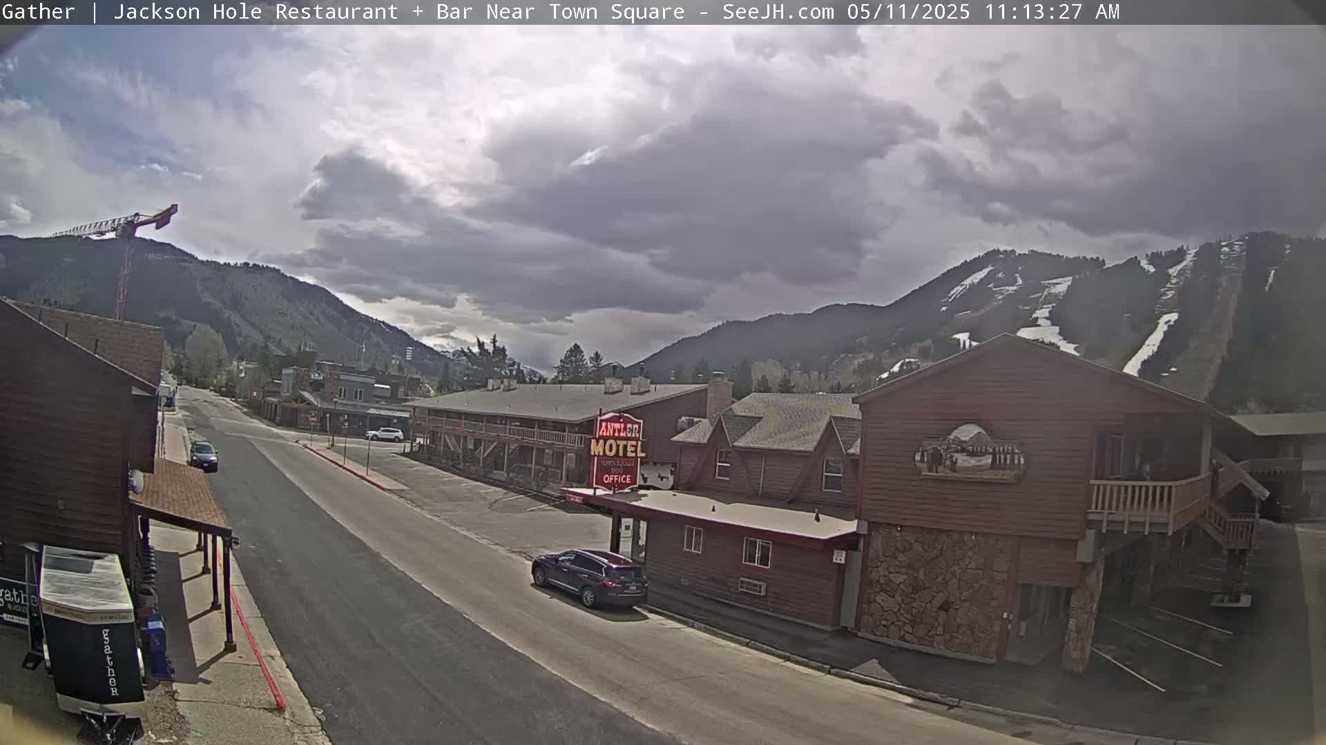 A mostly cloudy day reveals a mountain range with ski runs behind a street scene featuring a motel and a few vehicles.