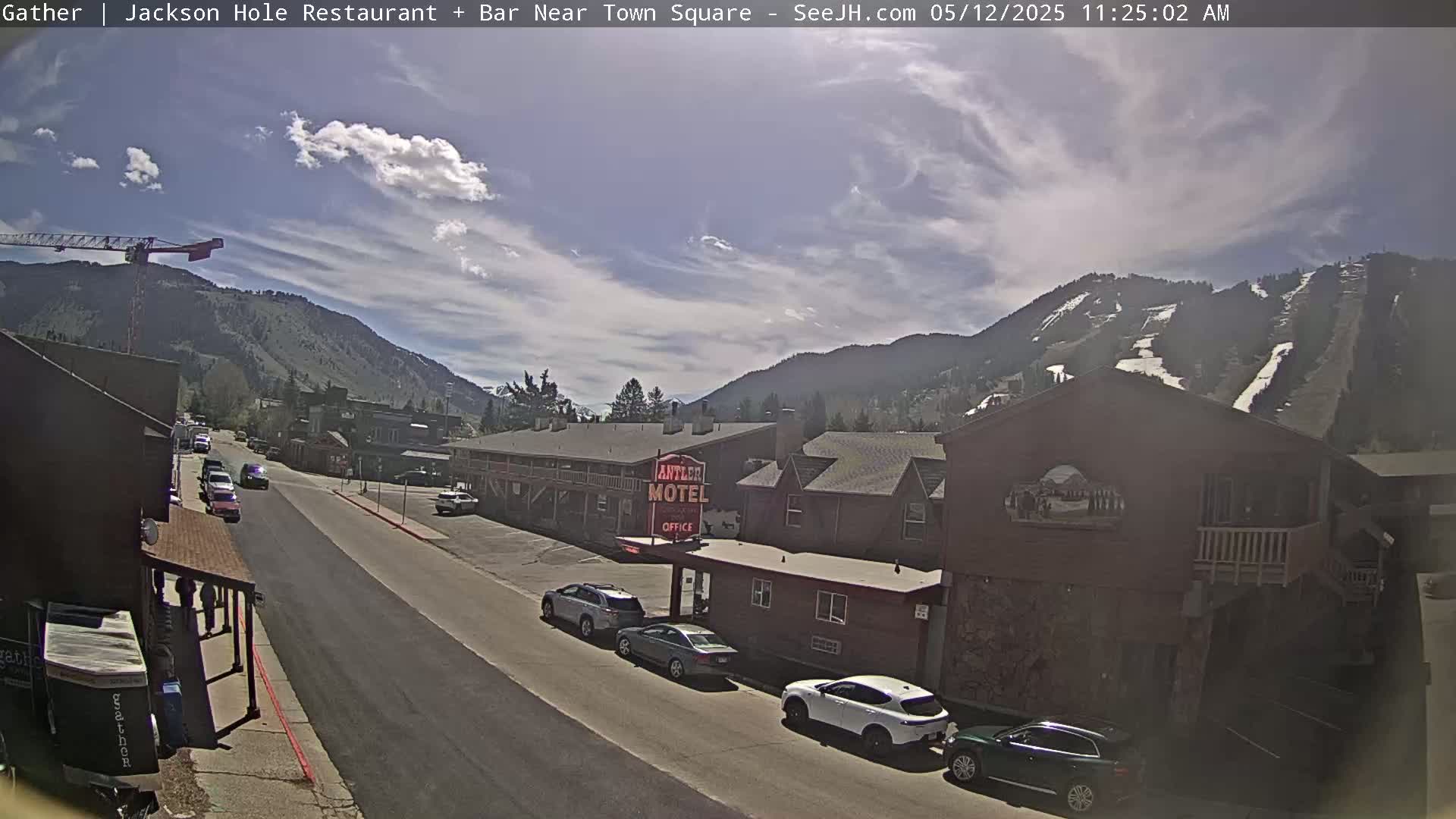 A partly cloudy day shows a street scene in a mountain town with cars parked along the side of a road,  a motel, and mountains in the background showing ski runs.
