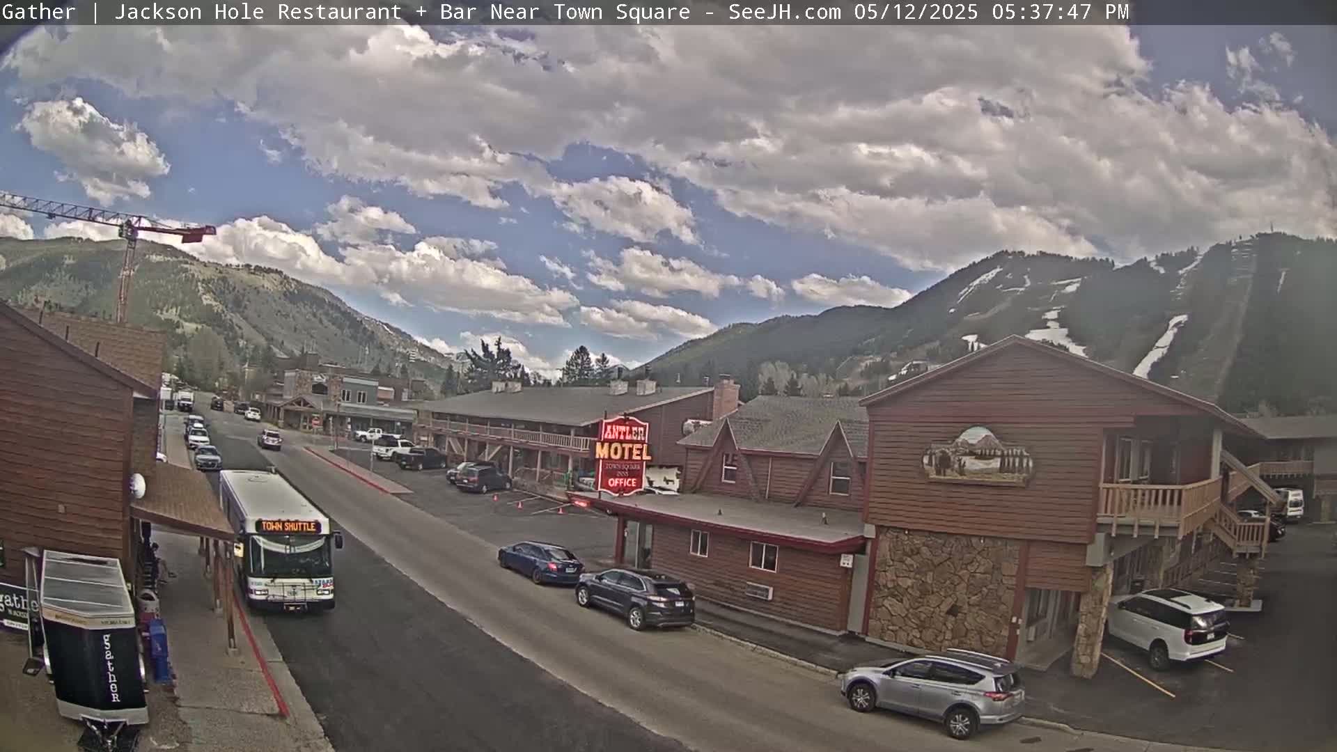 A partly cloudy day reveals a mountain town scene with cars parked along a street, a town shuttle bus, and a motel, all nestled against a backdrop of mountains.