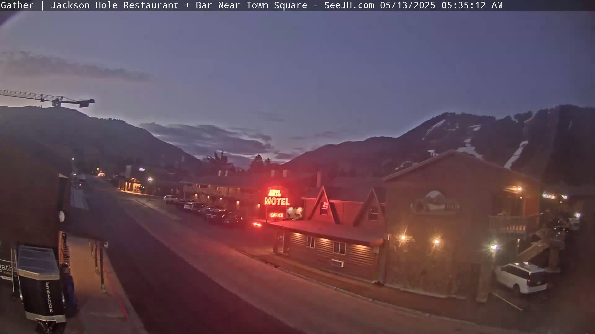 A dimly lit street scene at twilight shows a motel and other buildings nestled against a mountain range under a mostly clear sky.