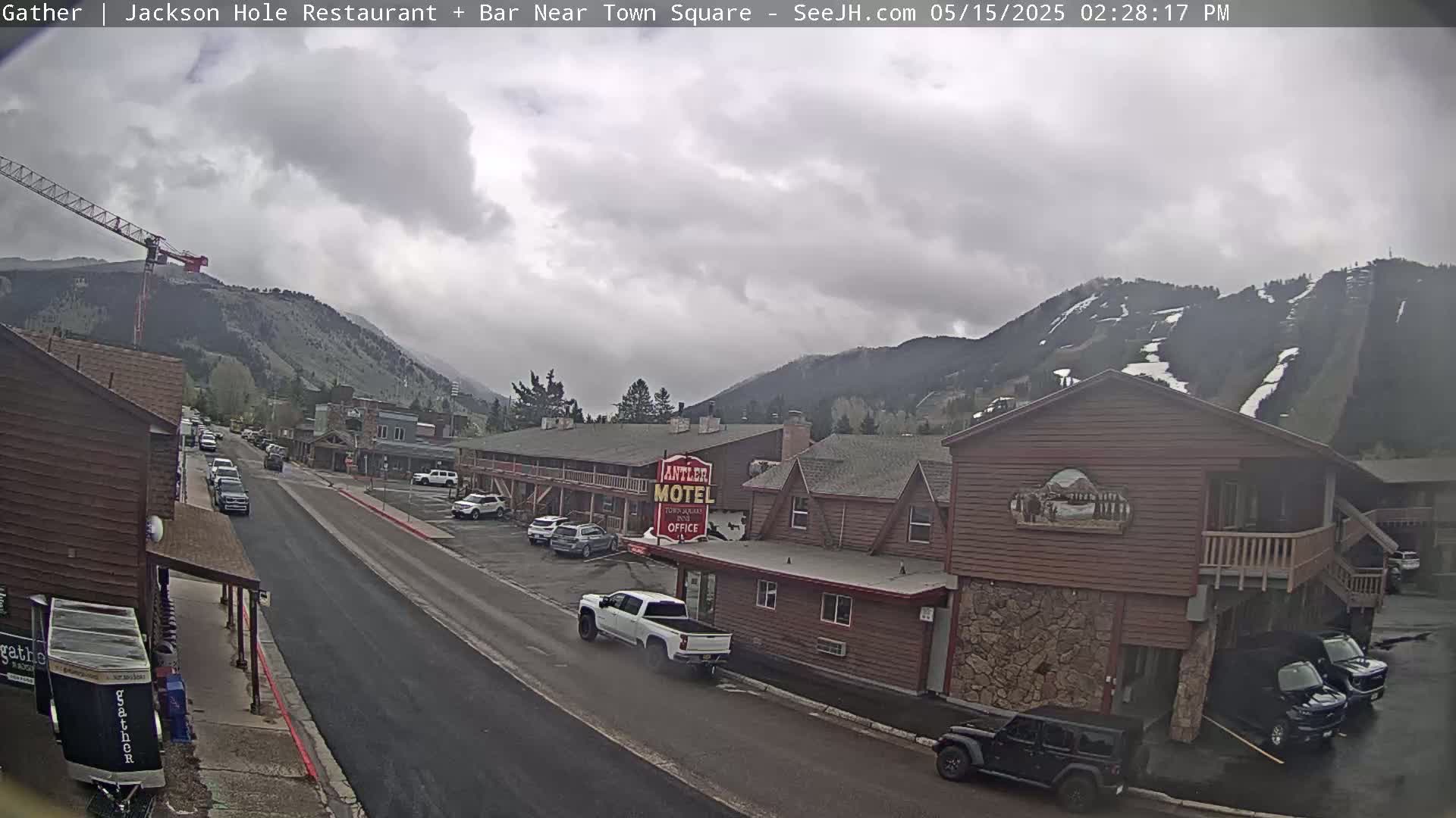 A cloudy day reveals a town street scene with parked cars, a motel, and mountains in the background, along which a construction crane is visible.