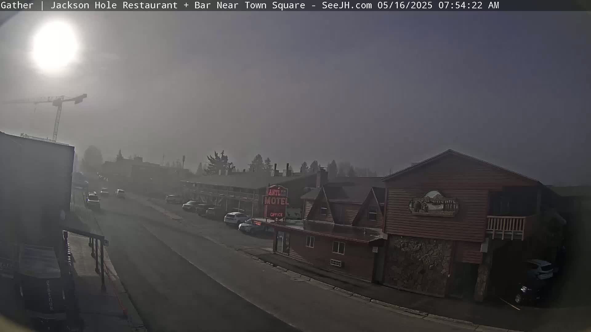 A foggy, sunlit street scene shows a motel and other buildings, with cars parked along the road and a construction crane visible in the distance.