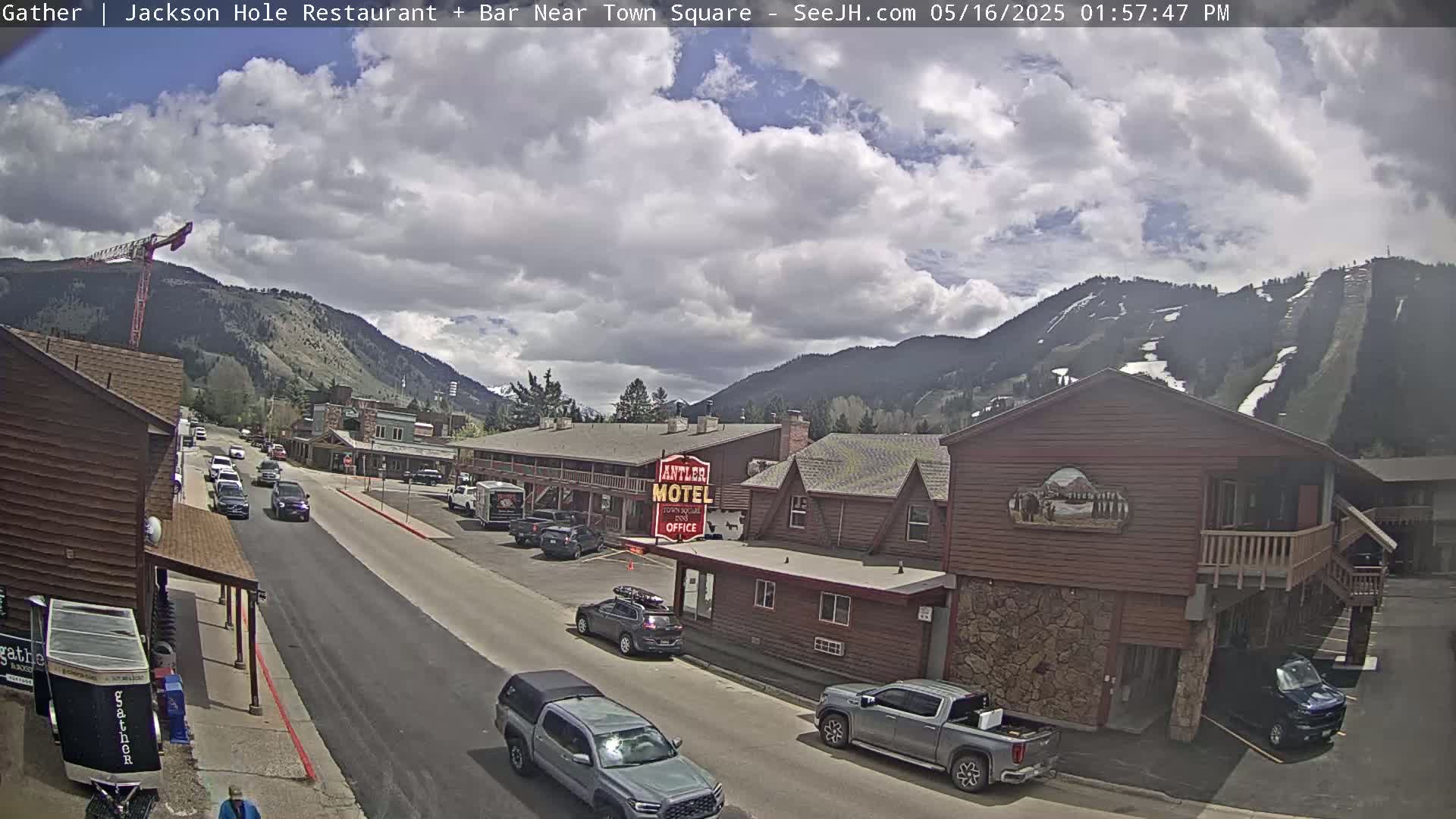 A partly cloudy day shows a street scene in a mountain town with cars parked and driving along a road, passing a motel and other buildings, with mountains and ski slopes in the background.