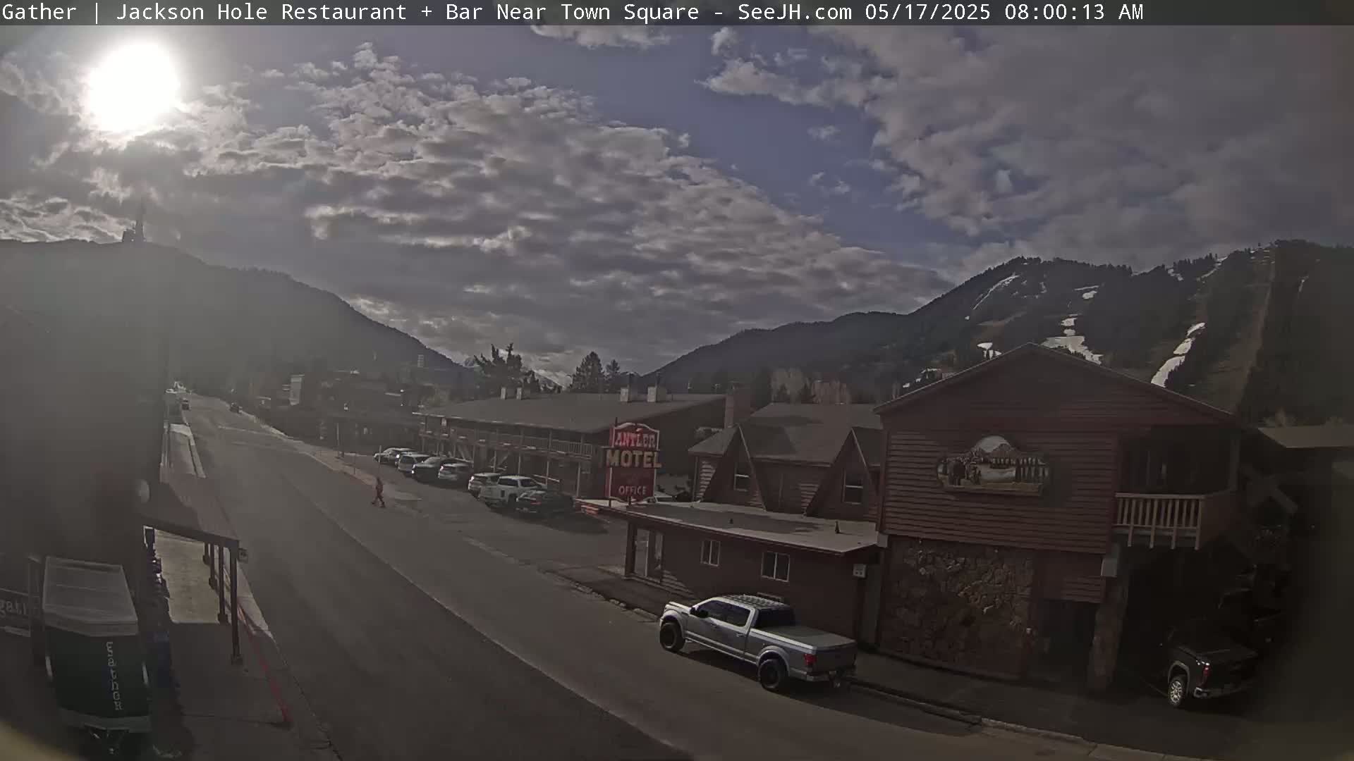A partly sunny day reveals a street scene with cars parked along the side of the road near a motel and other buildings, all nestled at the base of a mountain with ski runs visible on its slopes.