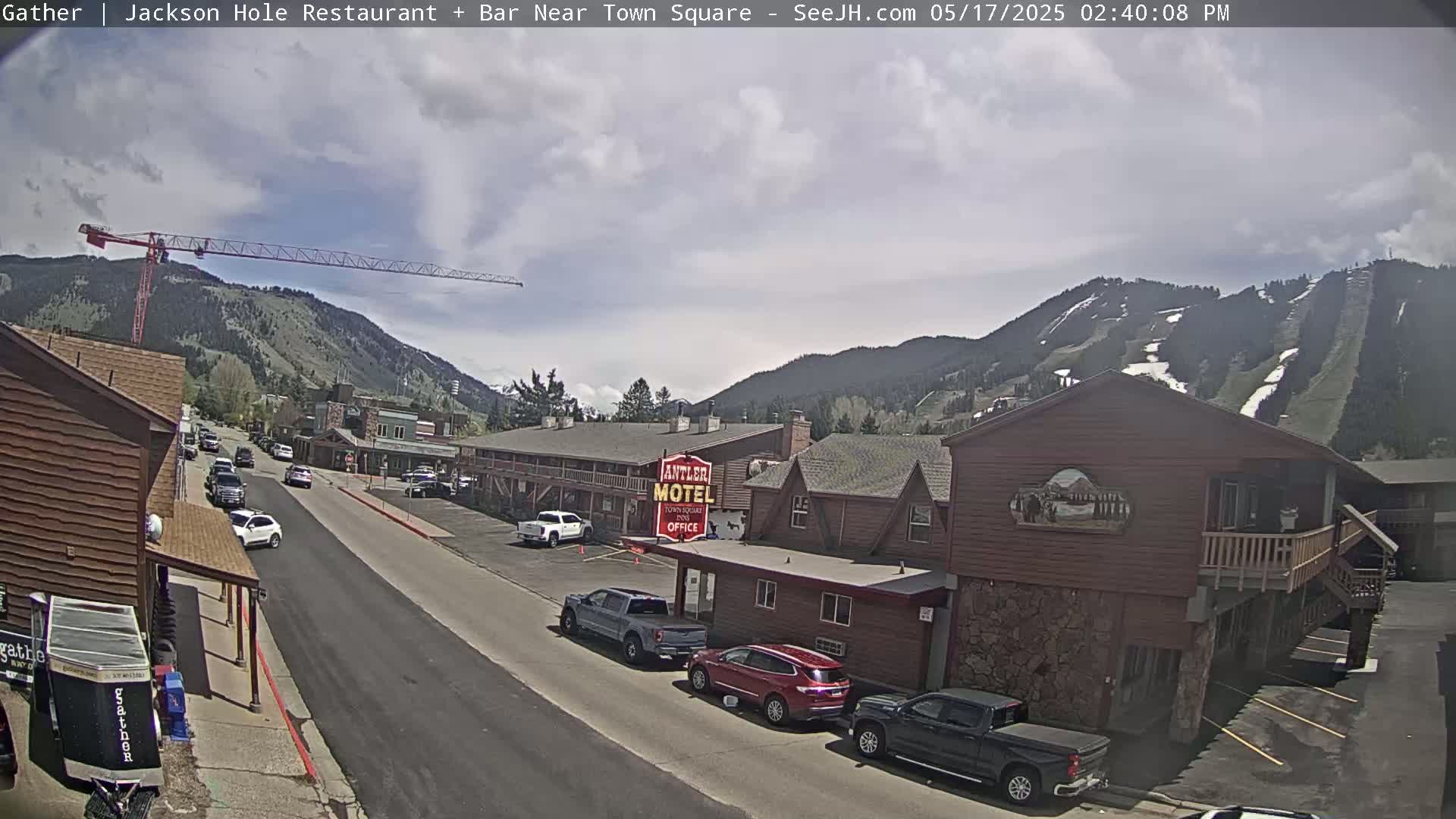 A partly cloudy day reveals a street scene with parked cars and buildings, including a motel, nestled against a mountain backdrop featuring visible ski slopes.