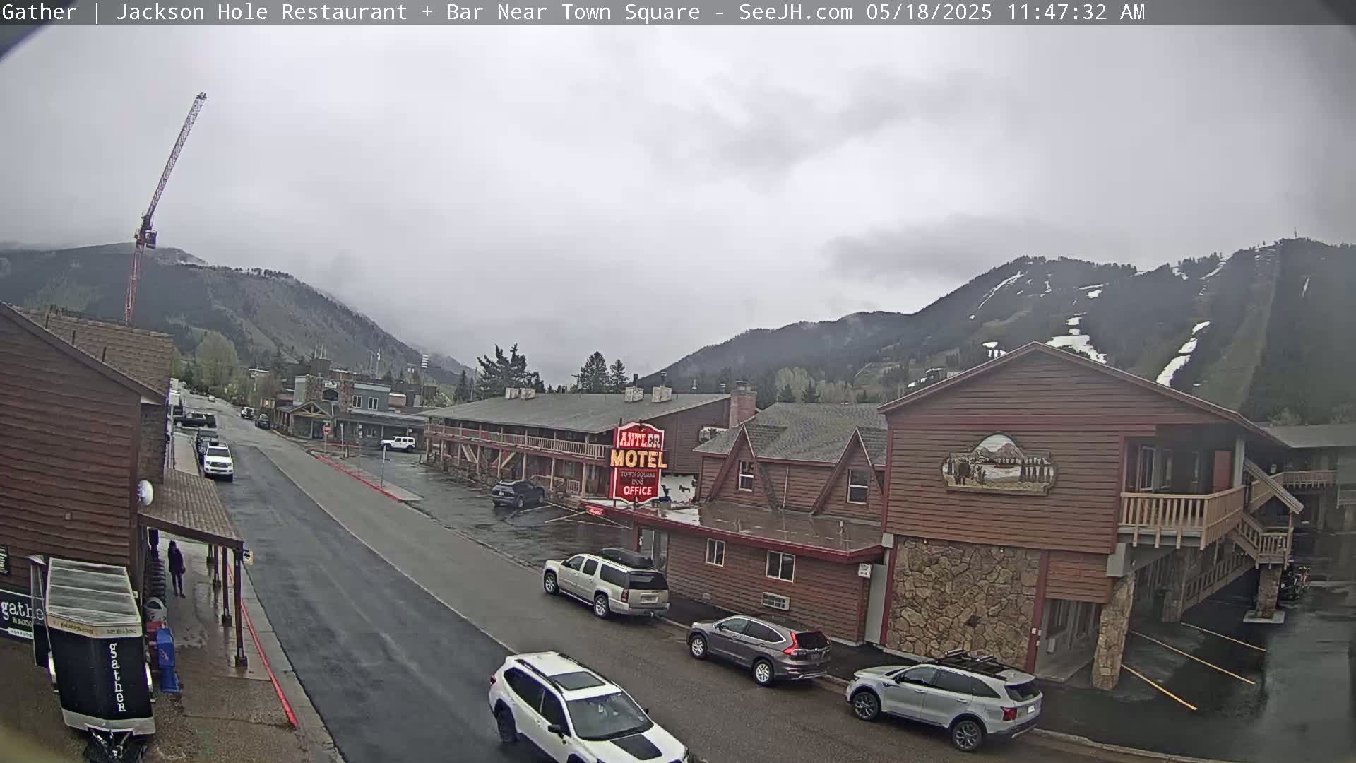 A wet street scene shows several cars parked in front of a wooden motel and other buildings, with overcast skies and mountains in the background.