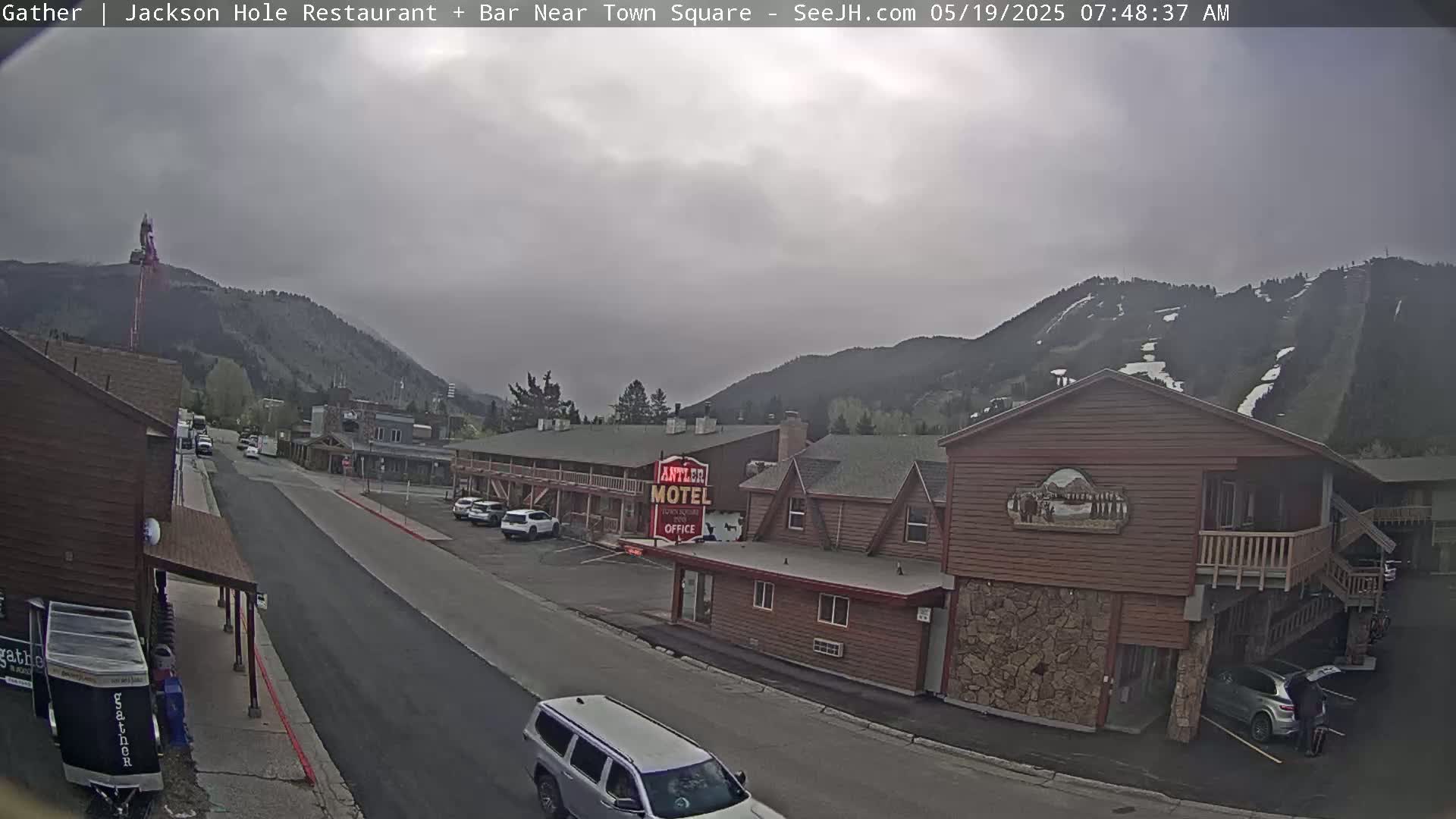 A street scene on an overcast day shows a motel and other buildings in a town nestled against a mountain range with visible ski slopes.