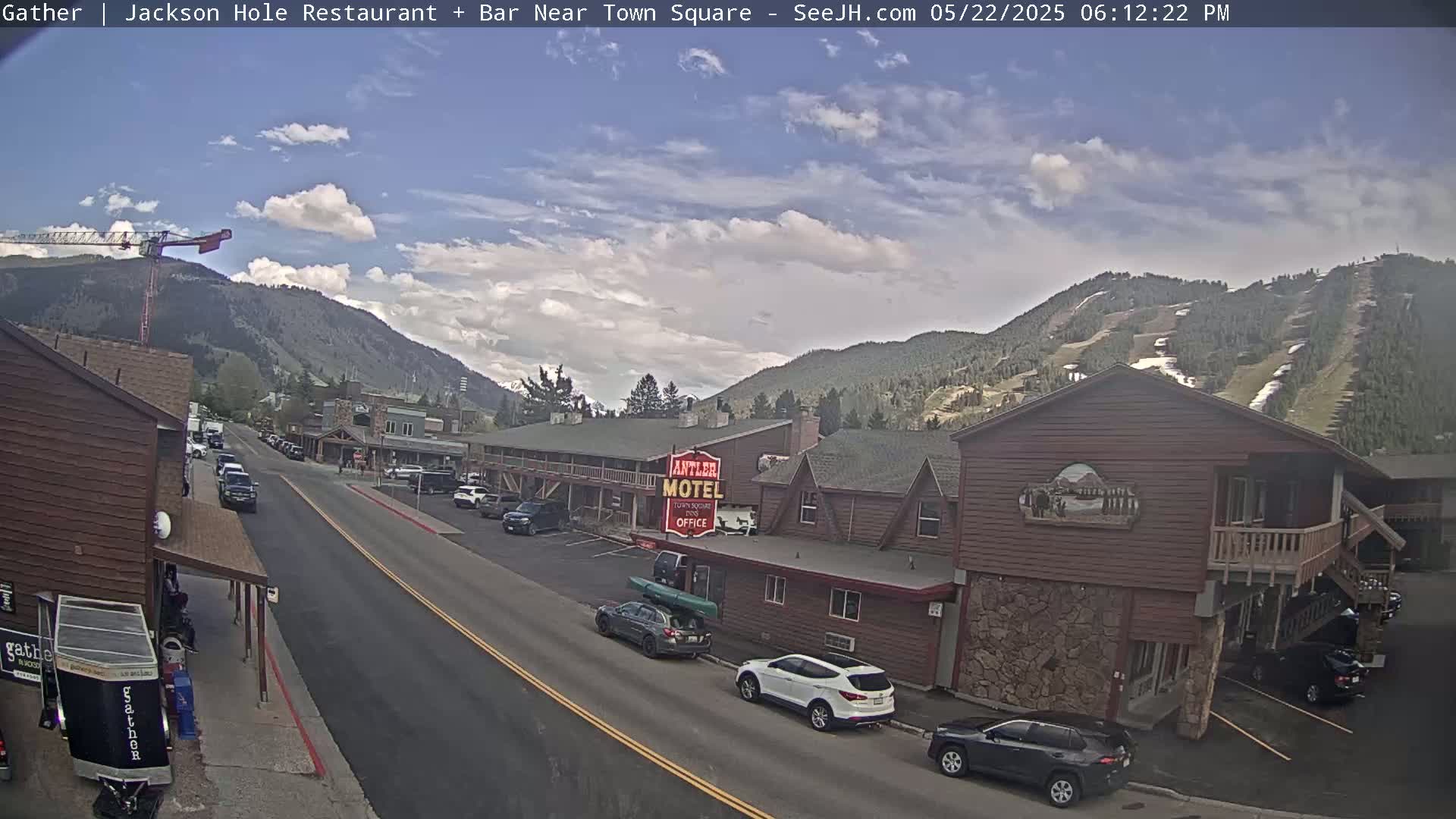 A partly cloudy day reveals a street scene with cars parked along the sides of a road in front of buildings, including a motel, set against a backdrop of mountains with visible ski slopes.
