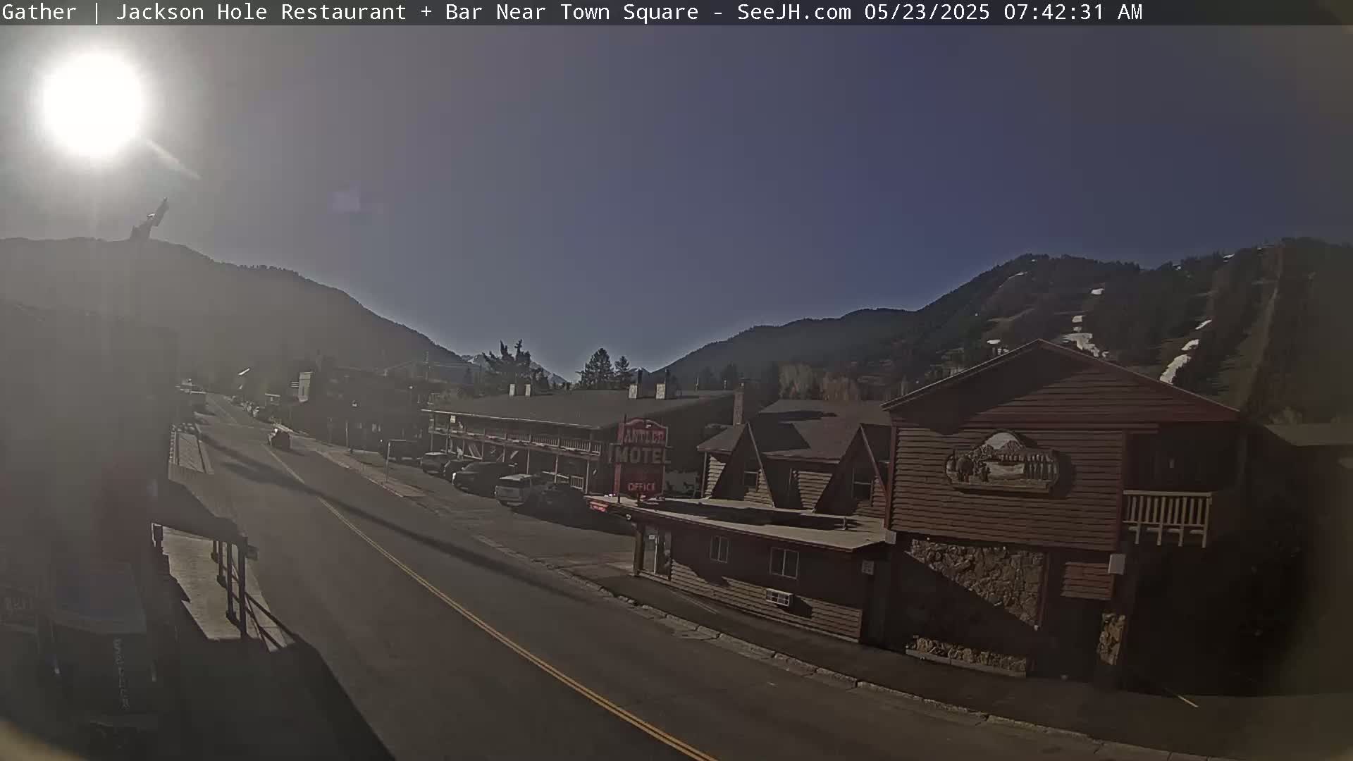 A sunny, clear day reveals a street scene with cars parked in front of a motel and other buildings nestled against a mountain backdrop.