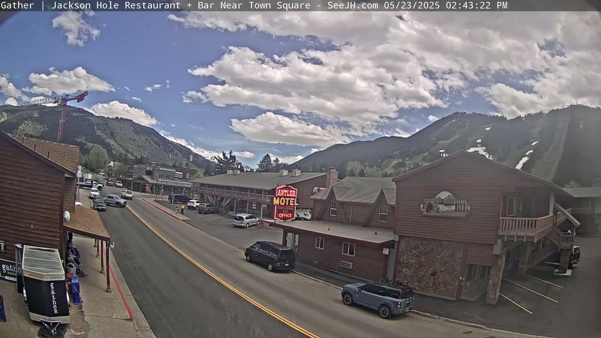 A partly cloudy day shows a street scene in a mountainous area with cars parked along the side of a road, in front of buildings including a motel.