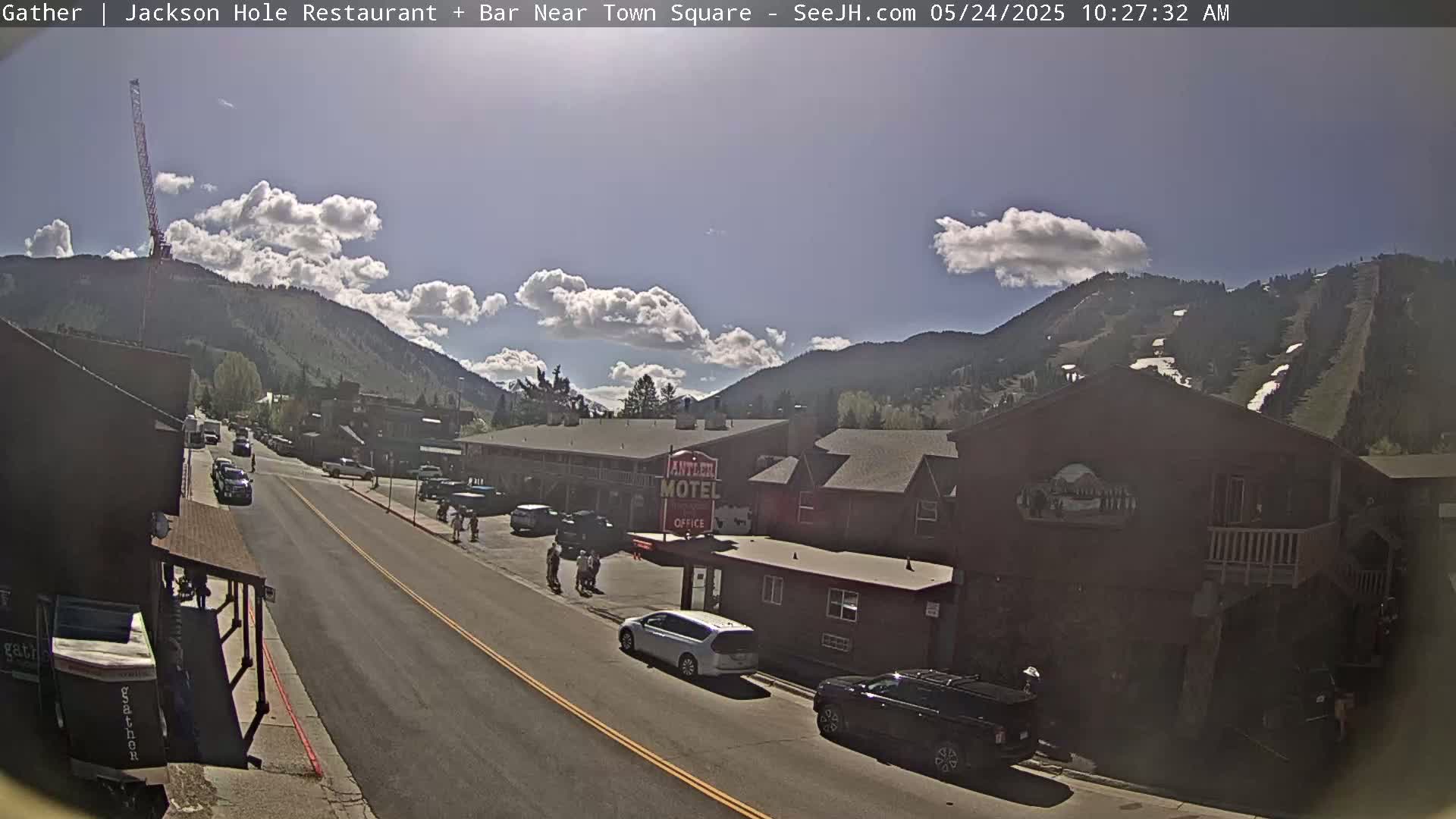 A sunny day with partly cloudy skies reveals a street scene in a mountain town, with cars parked along the sides of a road lined with buildings, including a motel.
