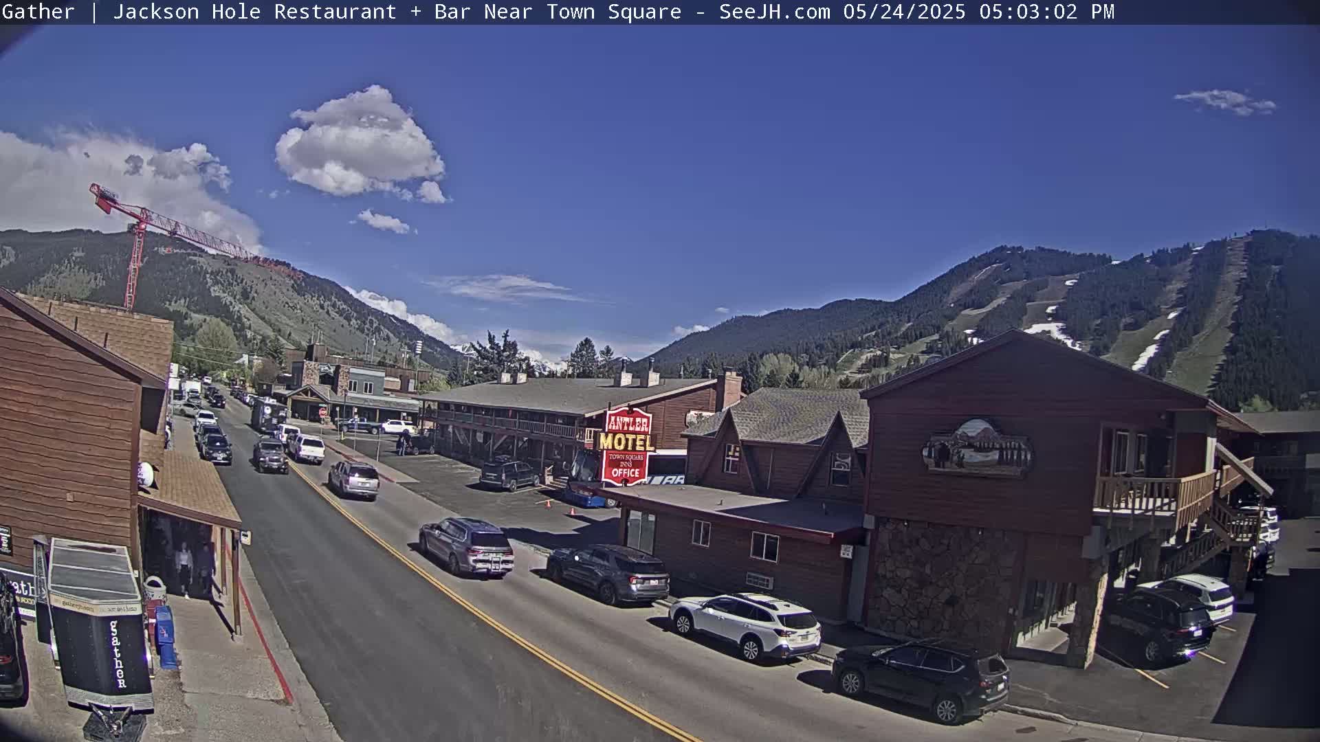 A partly sunny day shows a street scene in a mountain town with cars parked along the side of a road, and a motel and buildings in the background.