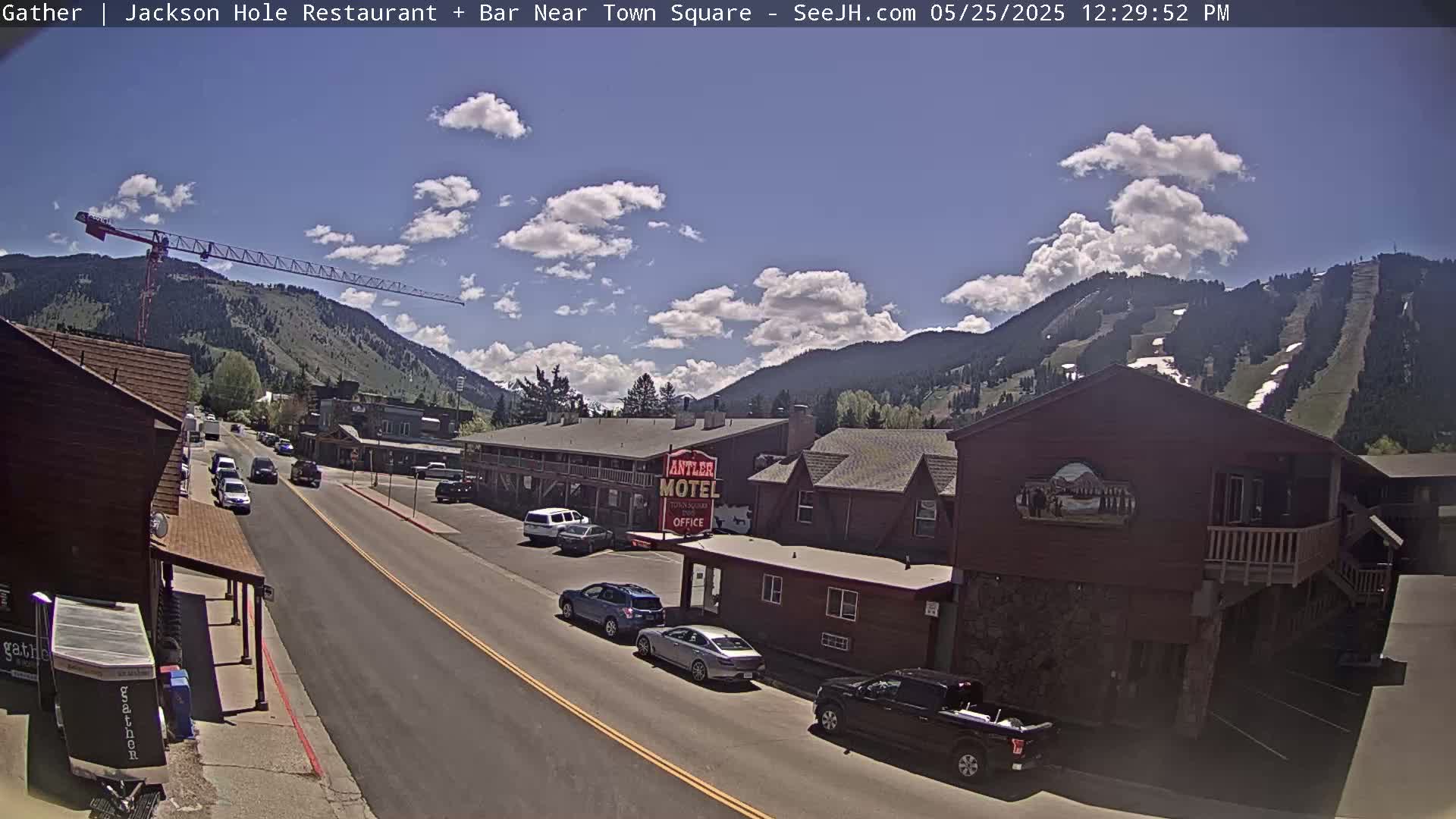 A sunny day reveals a town street scene with several cars parked and driving,  a motel, and a construction crane against a backdrop of mountains.