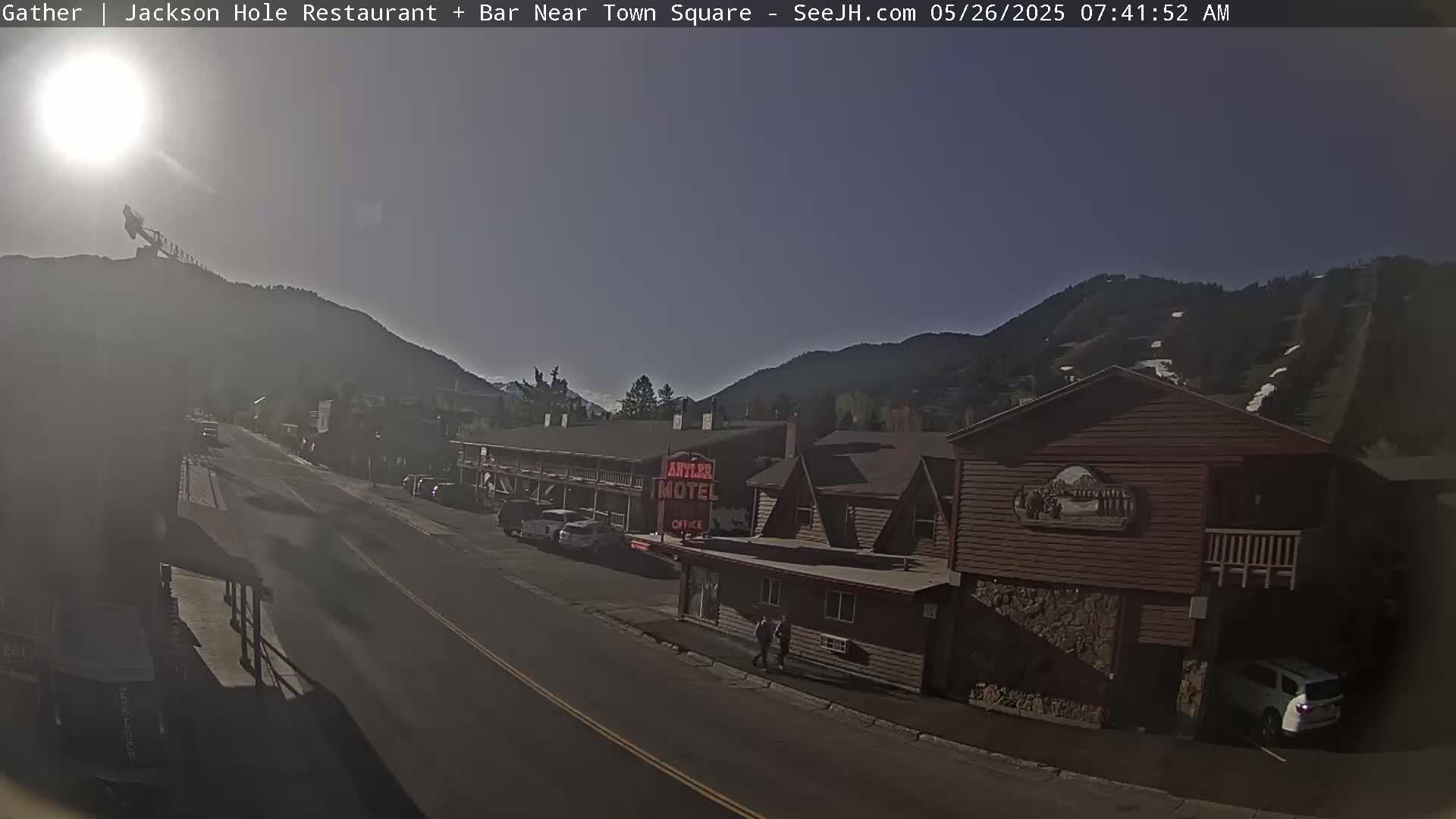 A sunny morning view of a street scene in a mountainous area, showing buildings, parked cars, and a motel.