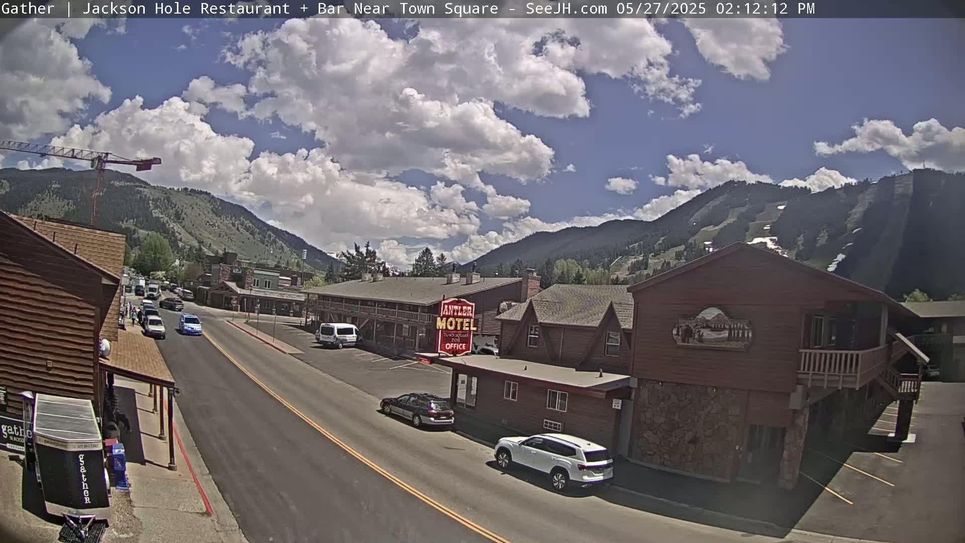 A partly sunny day reveals a street scene in a mountainous town, with cars parked along the roadside near a motel and buildings, under a partly cloudy sky.