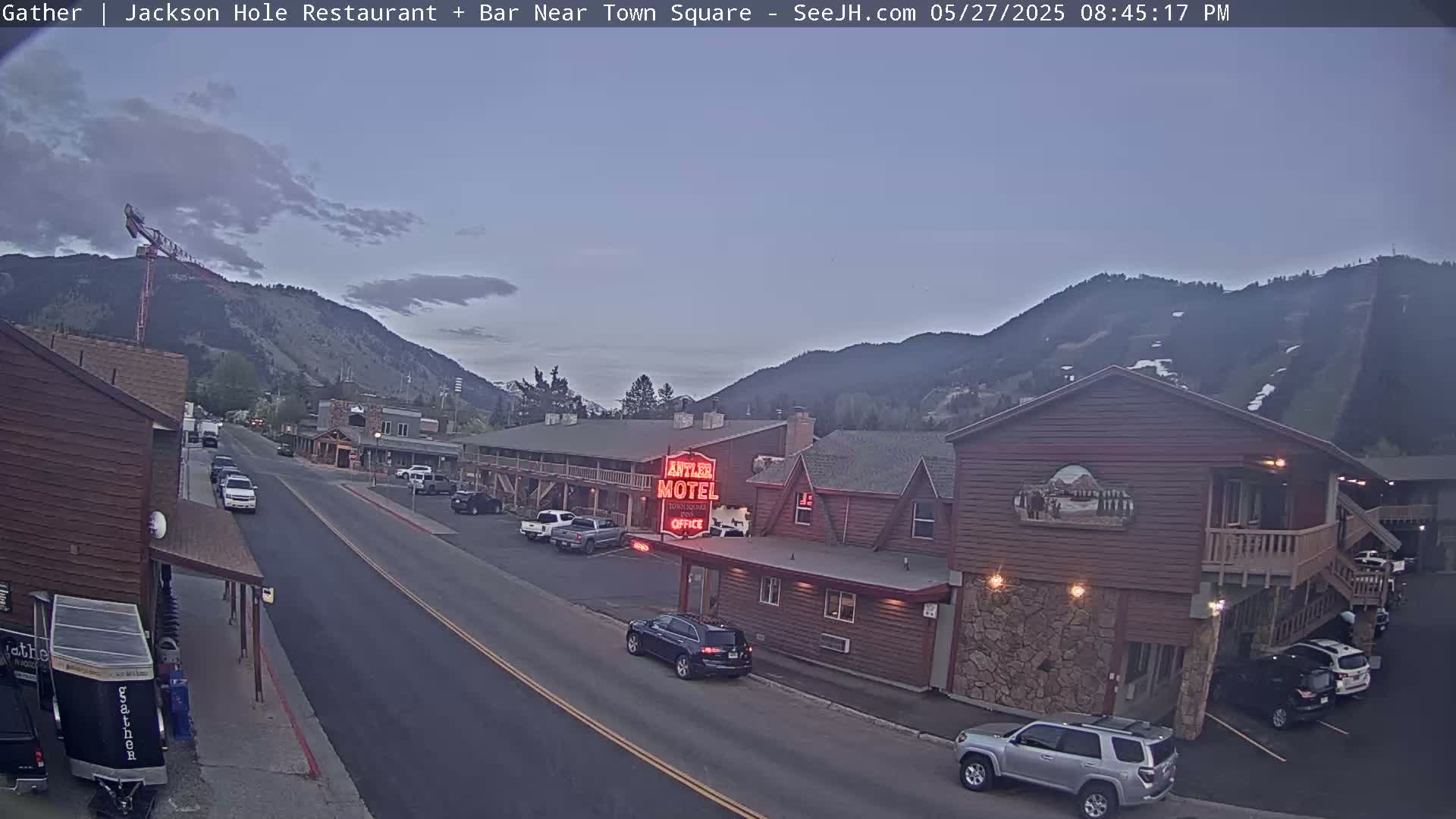 A street scene at dusk shows a motel and other buildings, cars parked along the street, and mountains in the background under a mostly clear sky.
