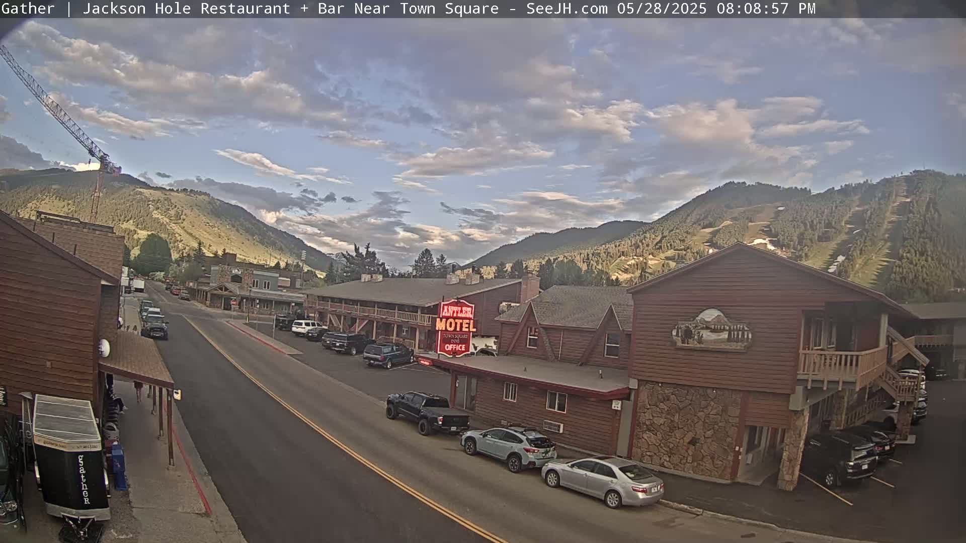 A partly cloudy afternoon scene shows a street with parked cars, leading to a motel and other buildings nestled at the base of a mountain with visible ski slopes.