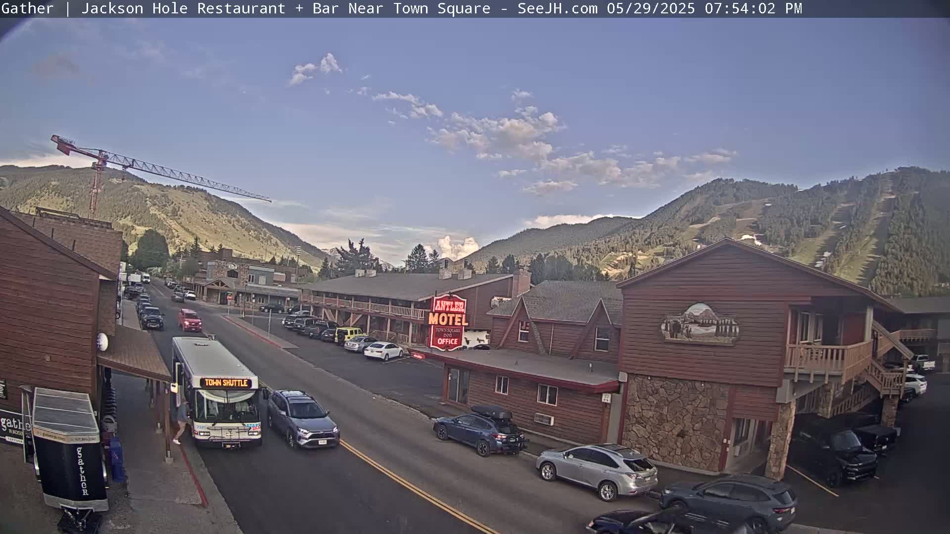 A partly cloudy afternoon scene shows a street with cars and a town shuttle bus in front of a motel and other buildings, with mountains in the background and a construction crane visible on a hillside.