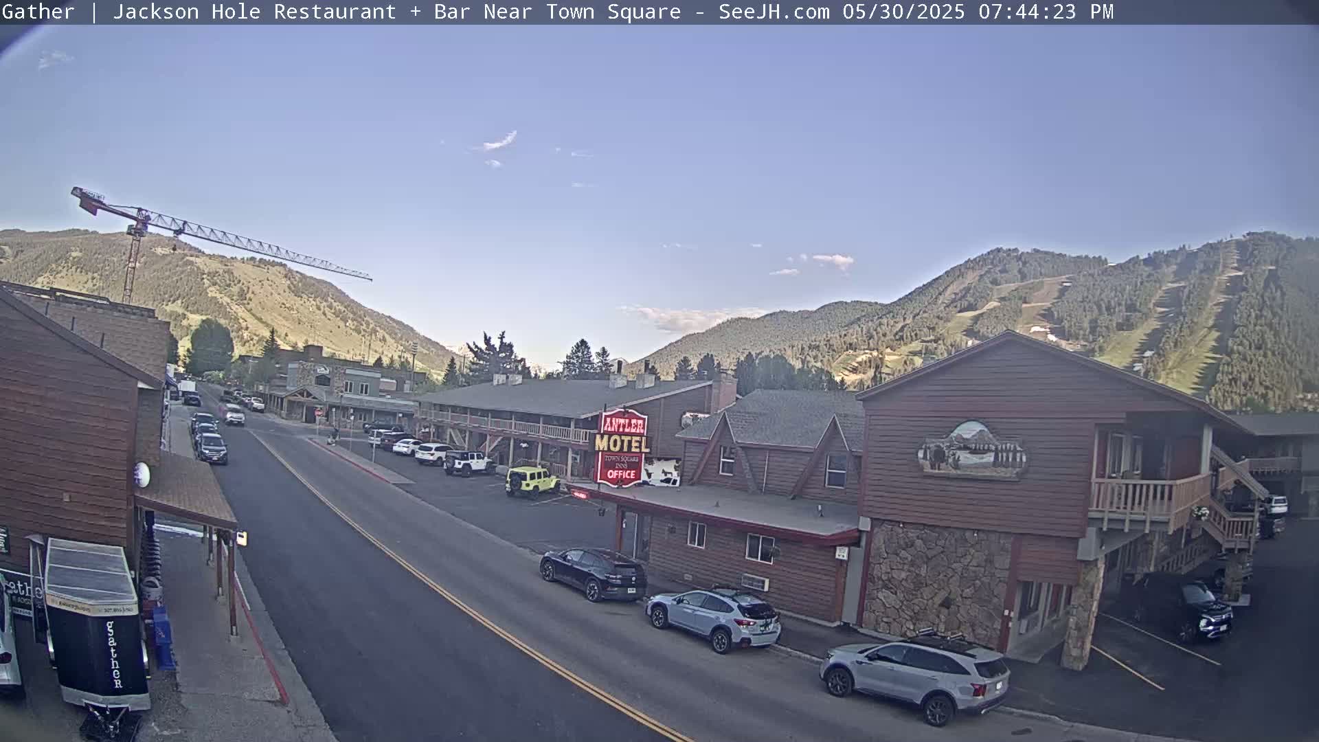 A sunny day reveals a town street scene nestled in a mountain valley, with cars parked along the roadside near a motel and a construction crane visible in the distance.